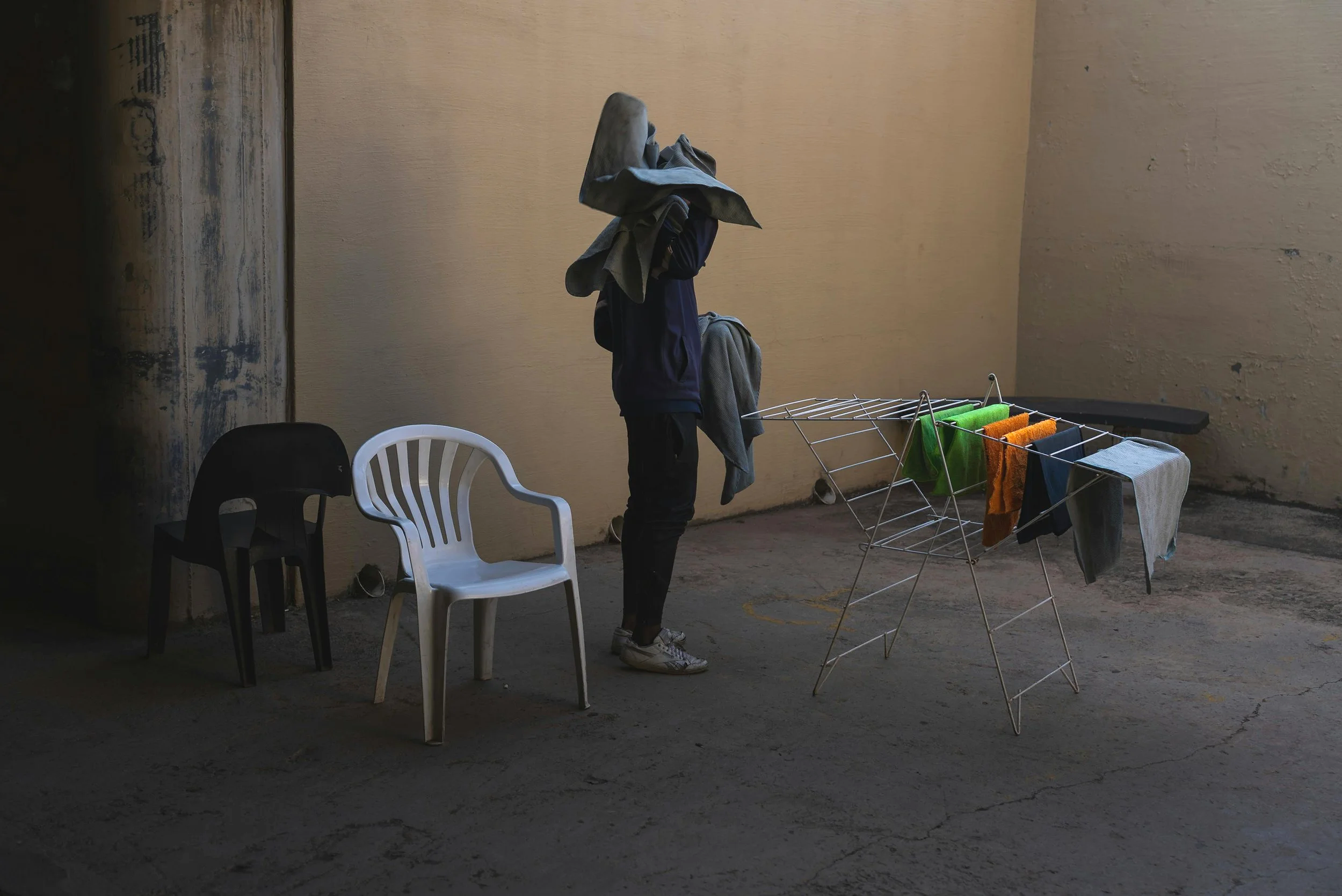 Drew Pollock standing outdoors next to a clothes rack with colourful towels hanging, with two chairs nearby, against a textured beige wall. A metaphor for finding the brand truth underneath organisational baggage.