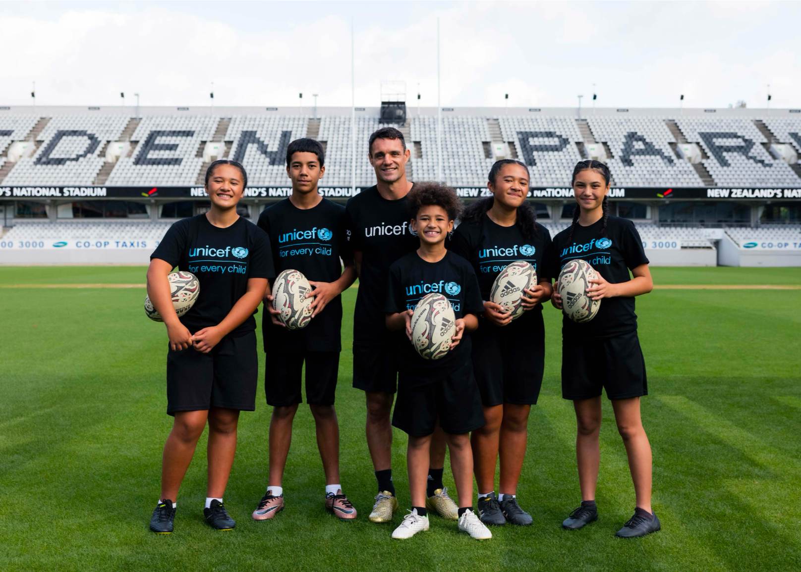 Beat Dan Carter campaign participants together on a stadium pitch holding rugby balls, showing the individual kicking idea becoming a shared participation experience.