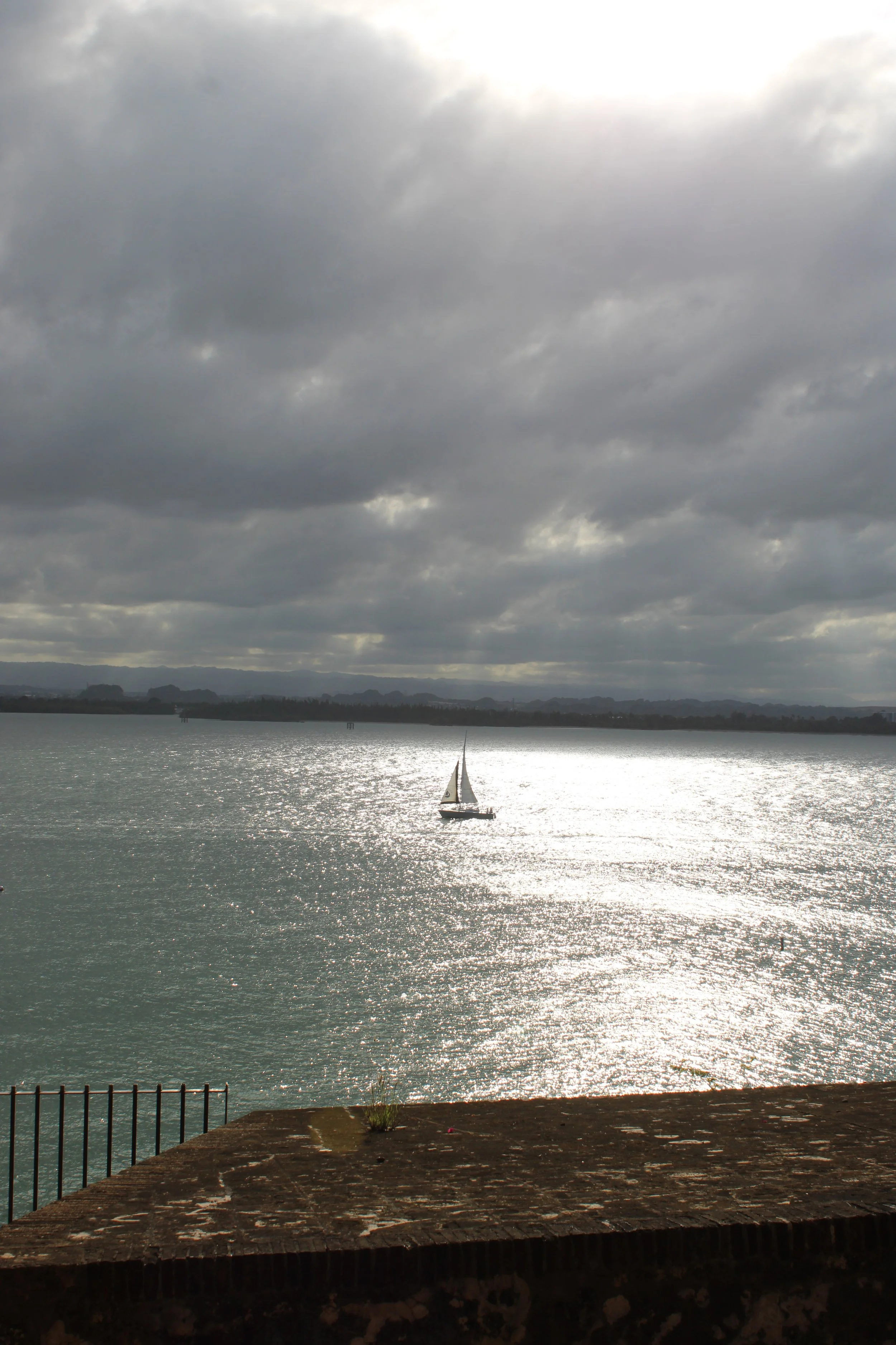 Overcast sky over a body of water with a sailboat in the distance and a stone patio with a metal railing in the foreground.
