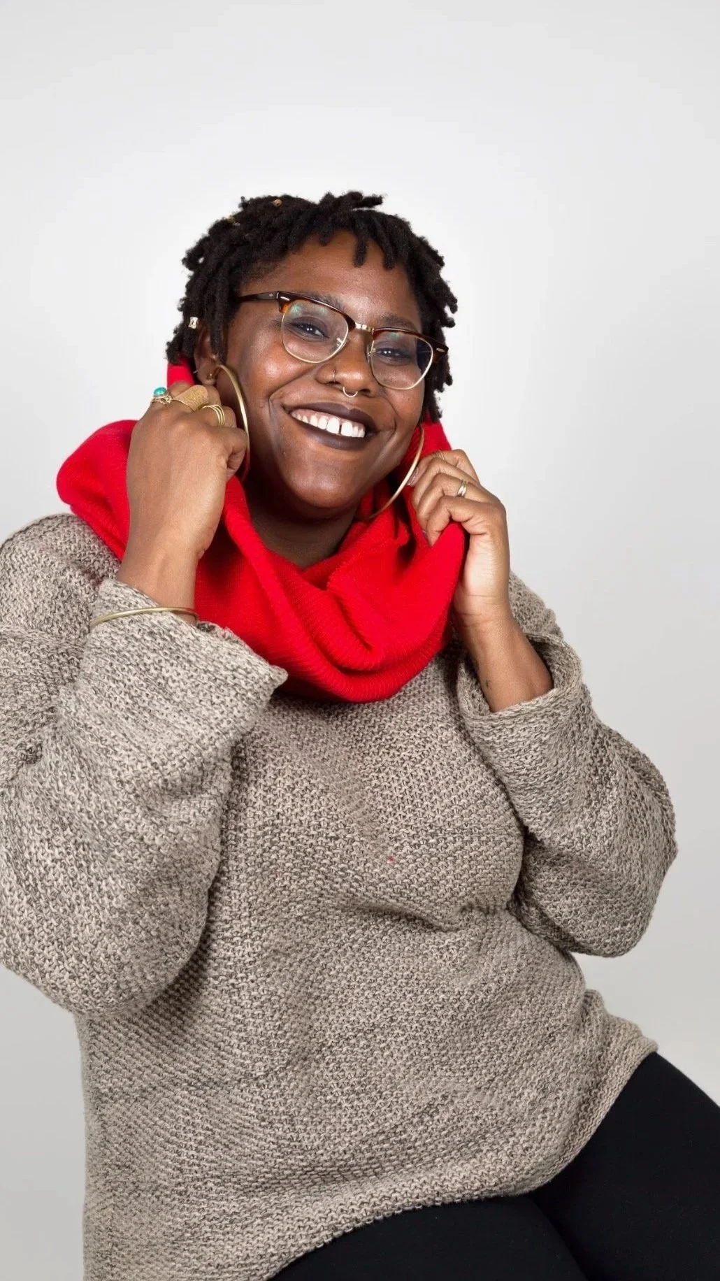 A woman with dreadlocks, glasses, and a septum piercing smiling and holding large hoop earrings in both hands. She is wearing a red scarf and a beige sweater, seated against a plain white background.