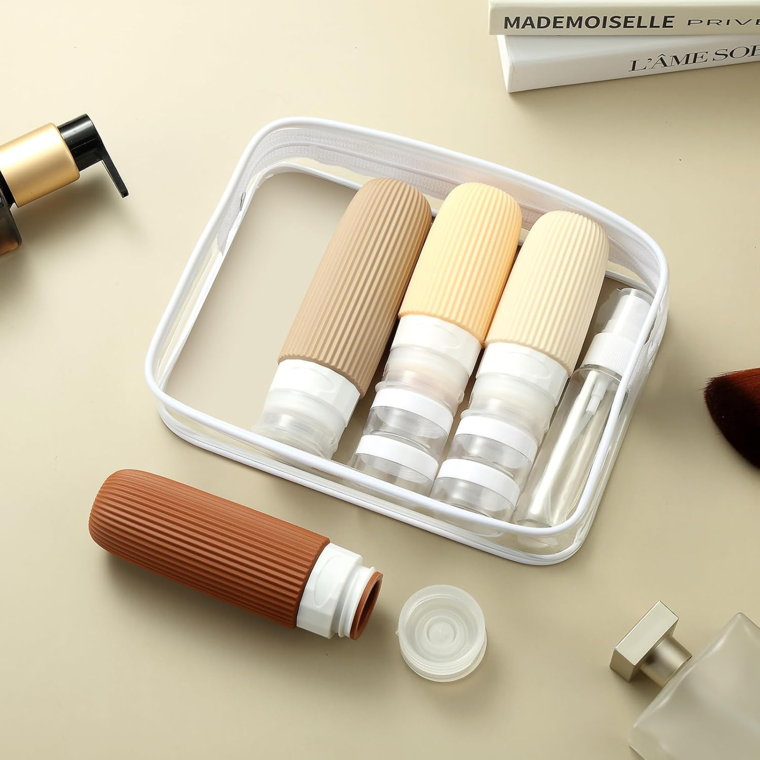 Clear travel bag holding three refillable squeeze bottles with ribbed neutral-tone caps, plus a small spray bottle.