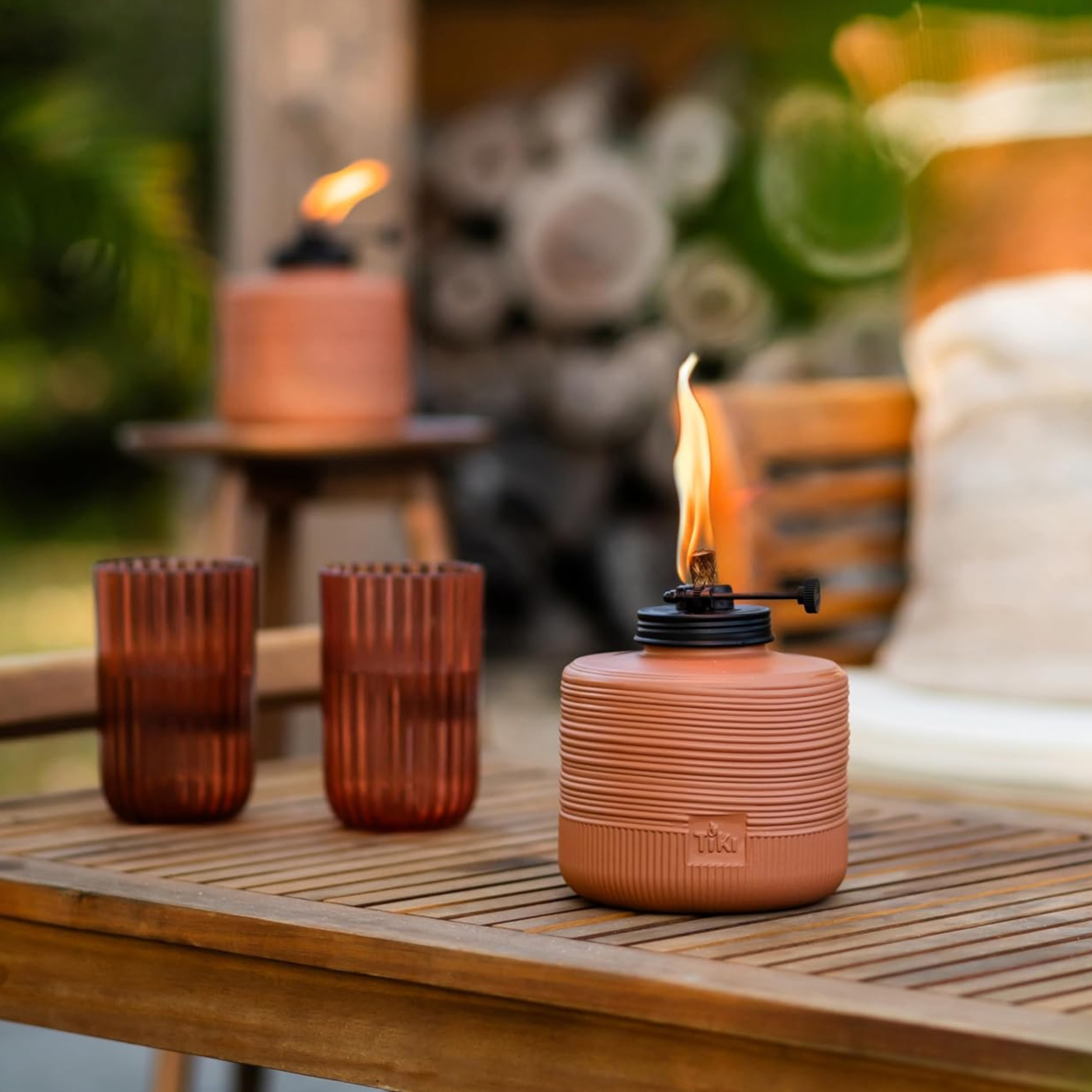 TIKI tabletop torch with live flame burning on a teak outdoor table beside two amber ribbed glass tumblers at golden hour with greenery in the background