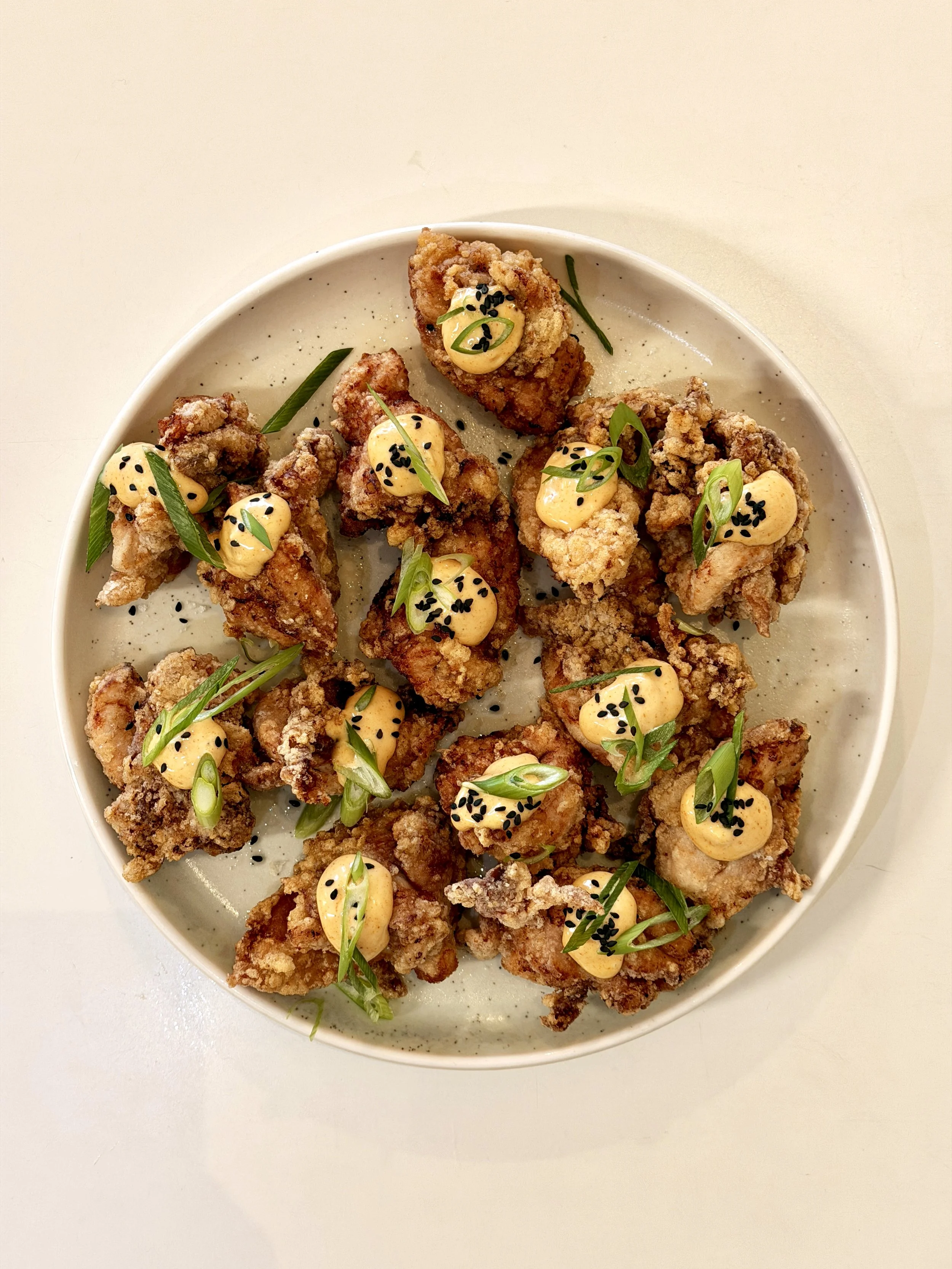 Fried chicken pieces topped with a creamy yellow sauce with black sesame seeds and chopped green onions on a white plate.