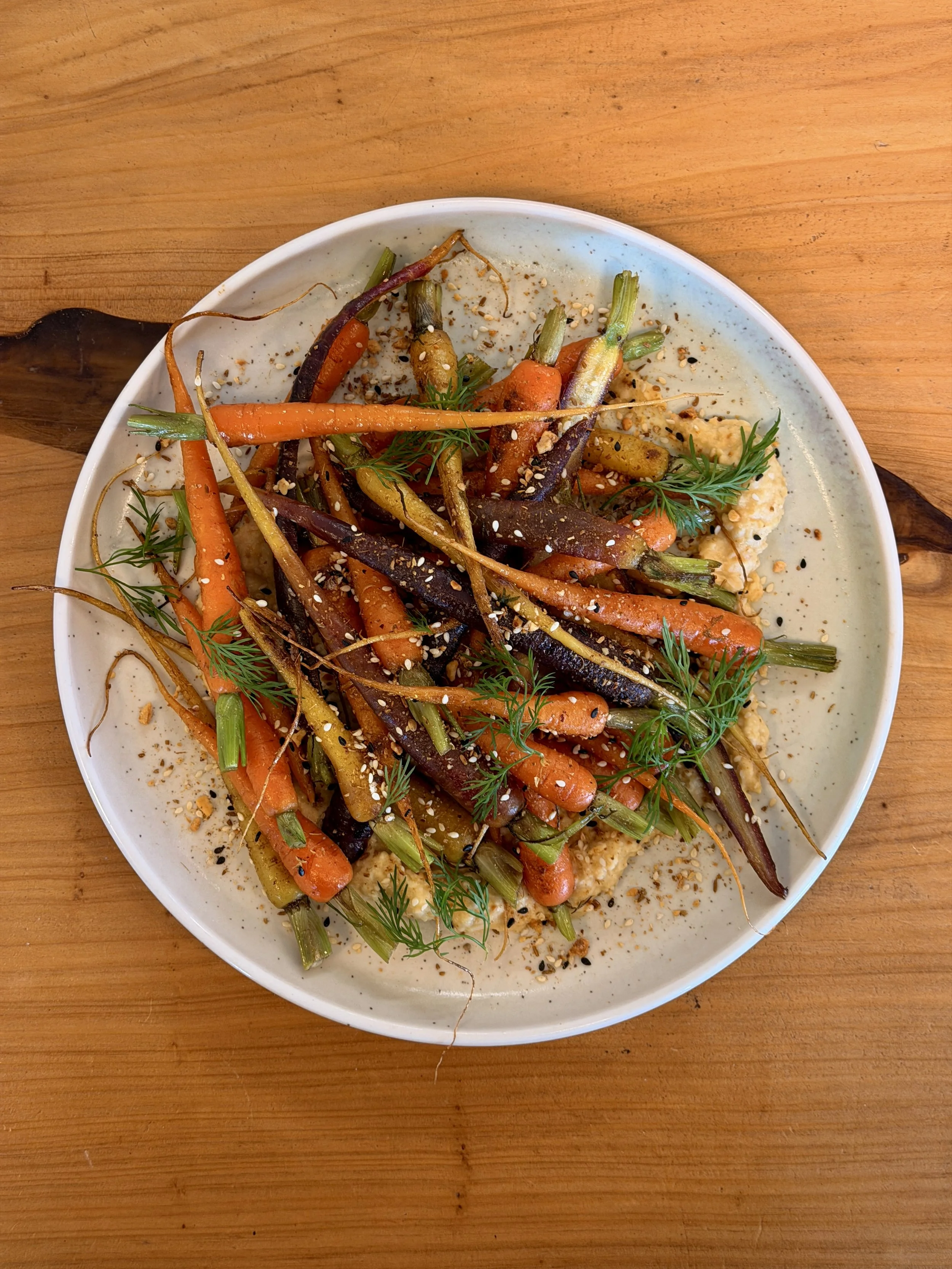 A white plate filled with roasted baby carrots garnished with fresh herbs, sesame seeds, and spices, placed on a wooden surface.