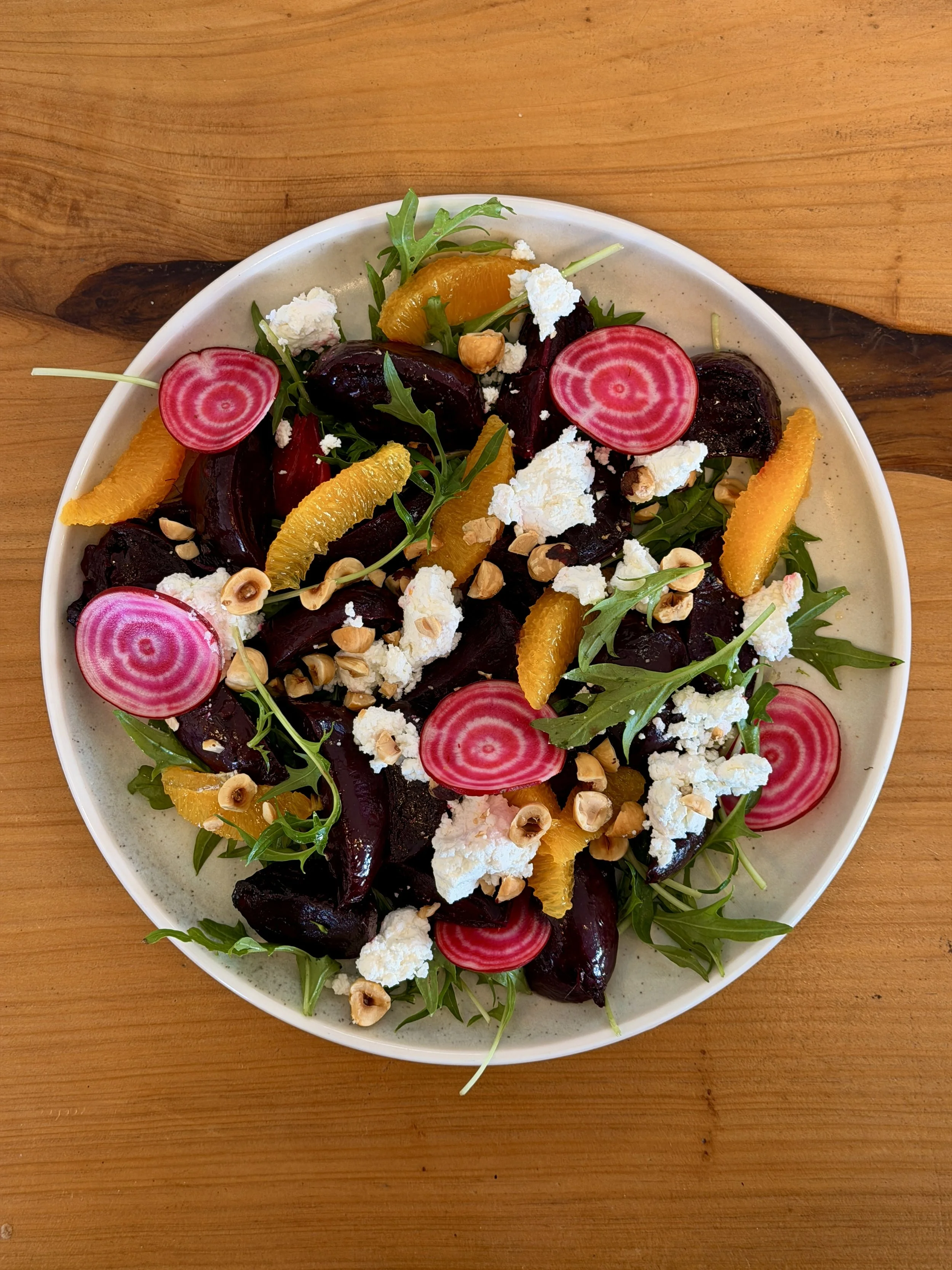 A salad with beets, orange slices, radish slices, arugula, crumbled cheese, chopped nuts, and microgreens in a white bowl on a wooden table.