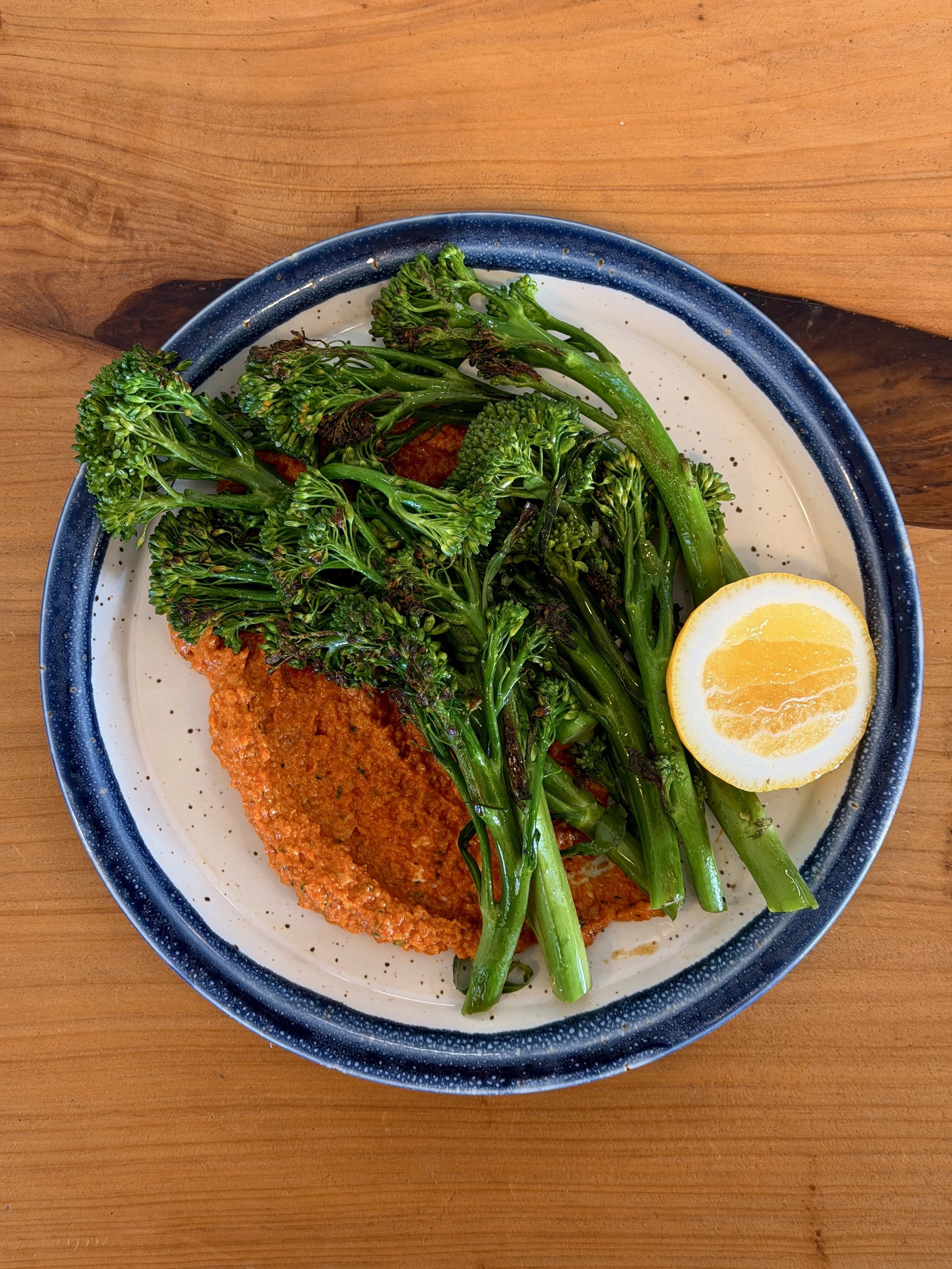 A plate of fried fish topped with broccolini and lemon wedge on a wooden table.