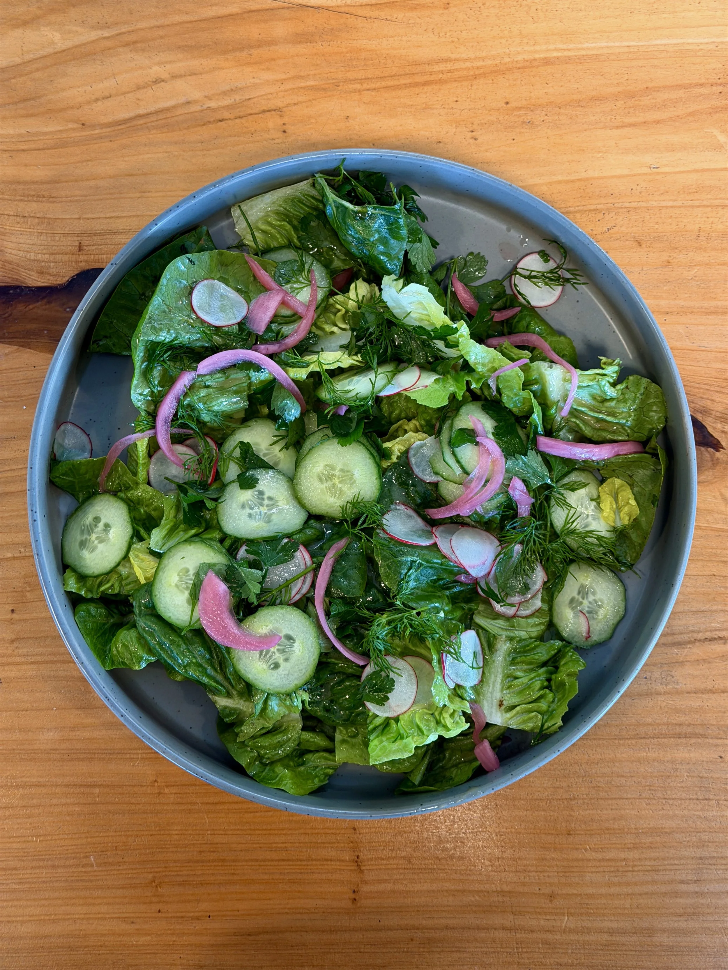 A bowl of fresh green salad containing lettuce, cucumber slices, radish slices, red onion slices, and herbs on a wooden table.