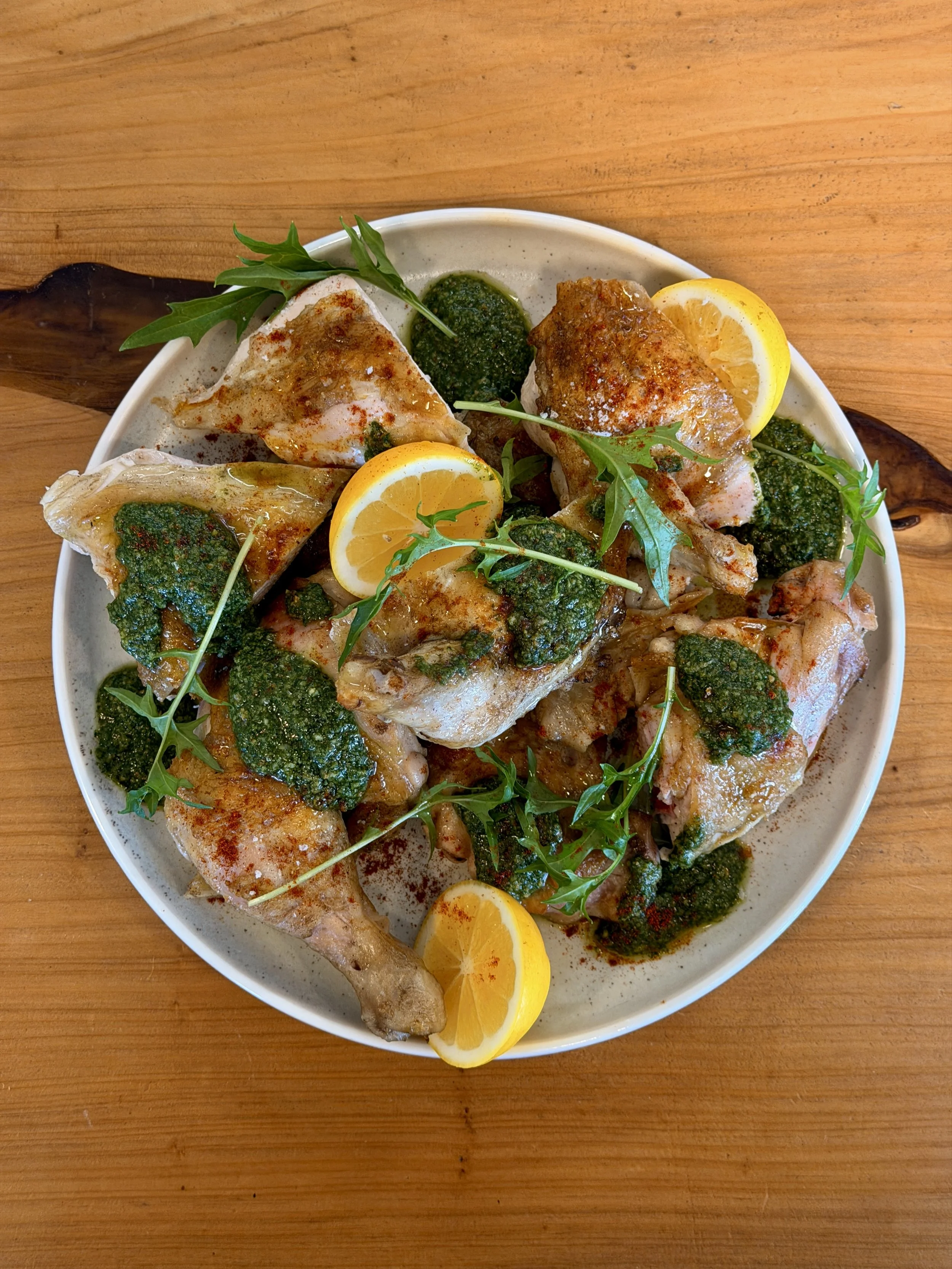 A white plate with cooked fish topped with green sauce, garnished with lemon slices and fresh green herbs, on a wooden table.