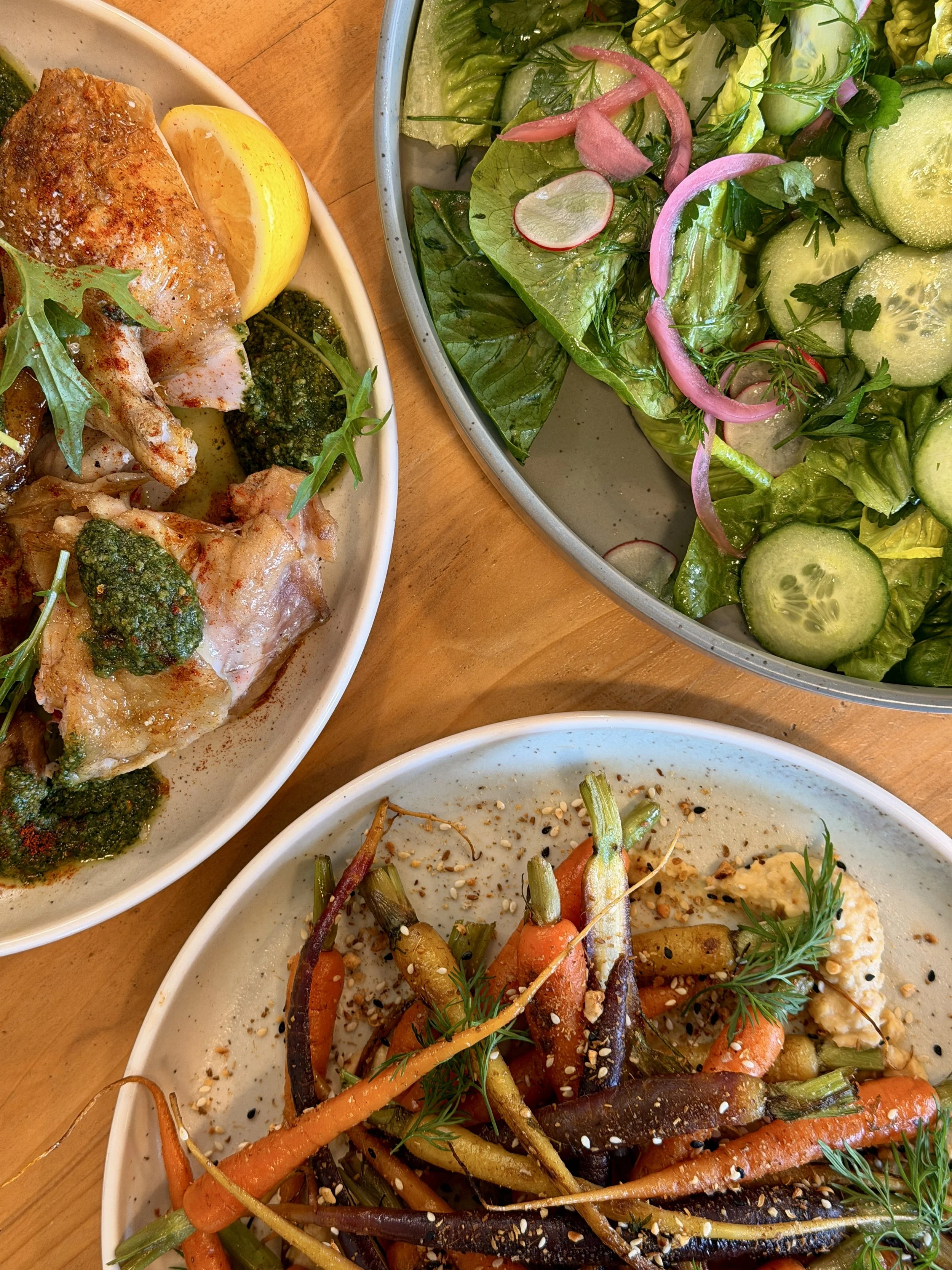 Close-up view of three plates of food on a wooden table, including a salad with cucumbers, radishes, and pink pickled onions, a roasted chicken leg with lemon and green sauce, and a dish with roasted carrots and other vegetables garnished with herbs.