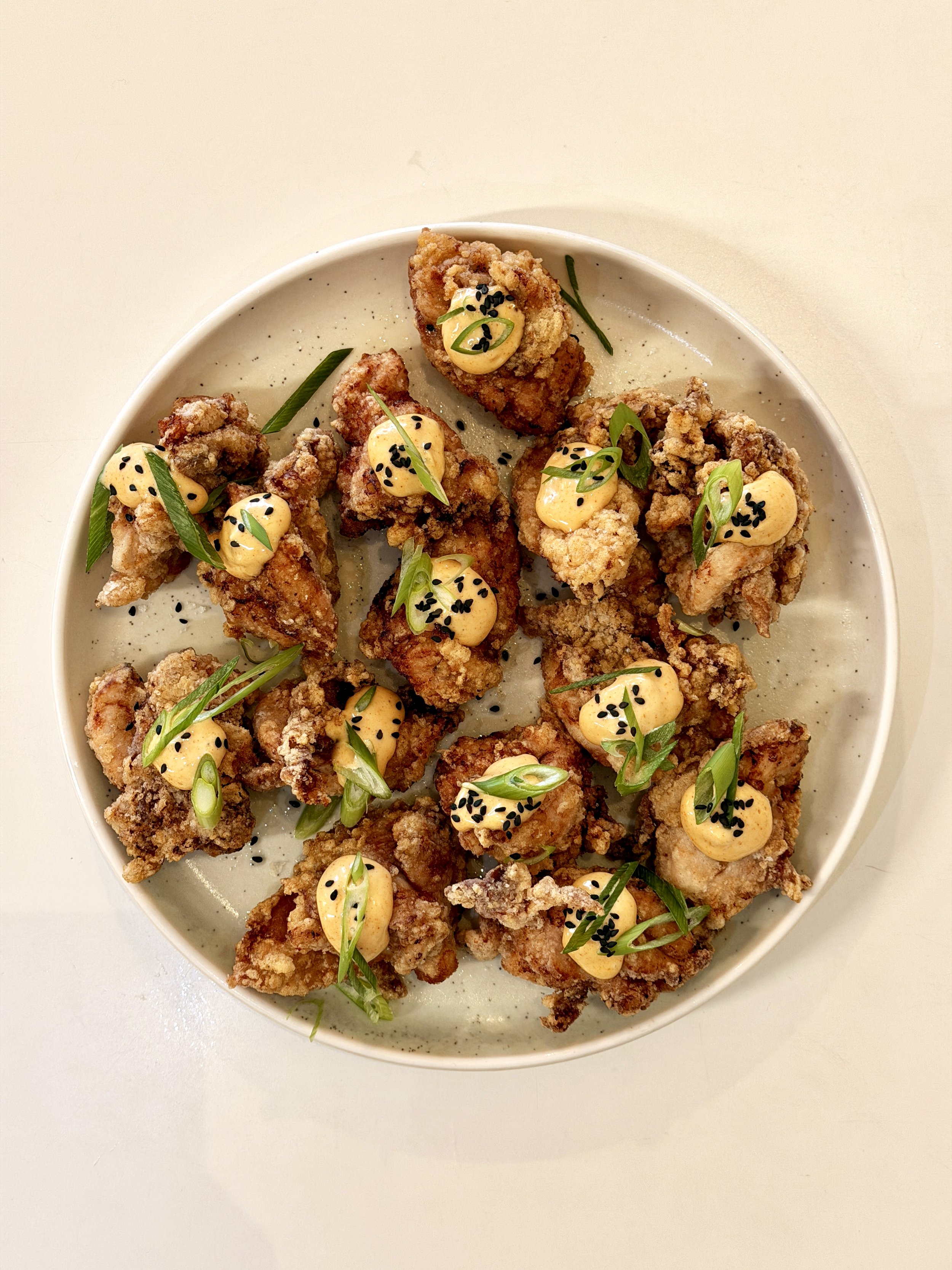 Fried chicken pieces topped with a creamy yellow sauce, black sesame seeds, and garnished with chopped green onions on a round ceramic plate.