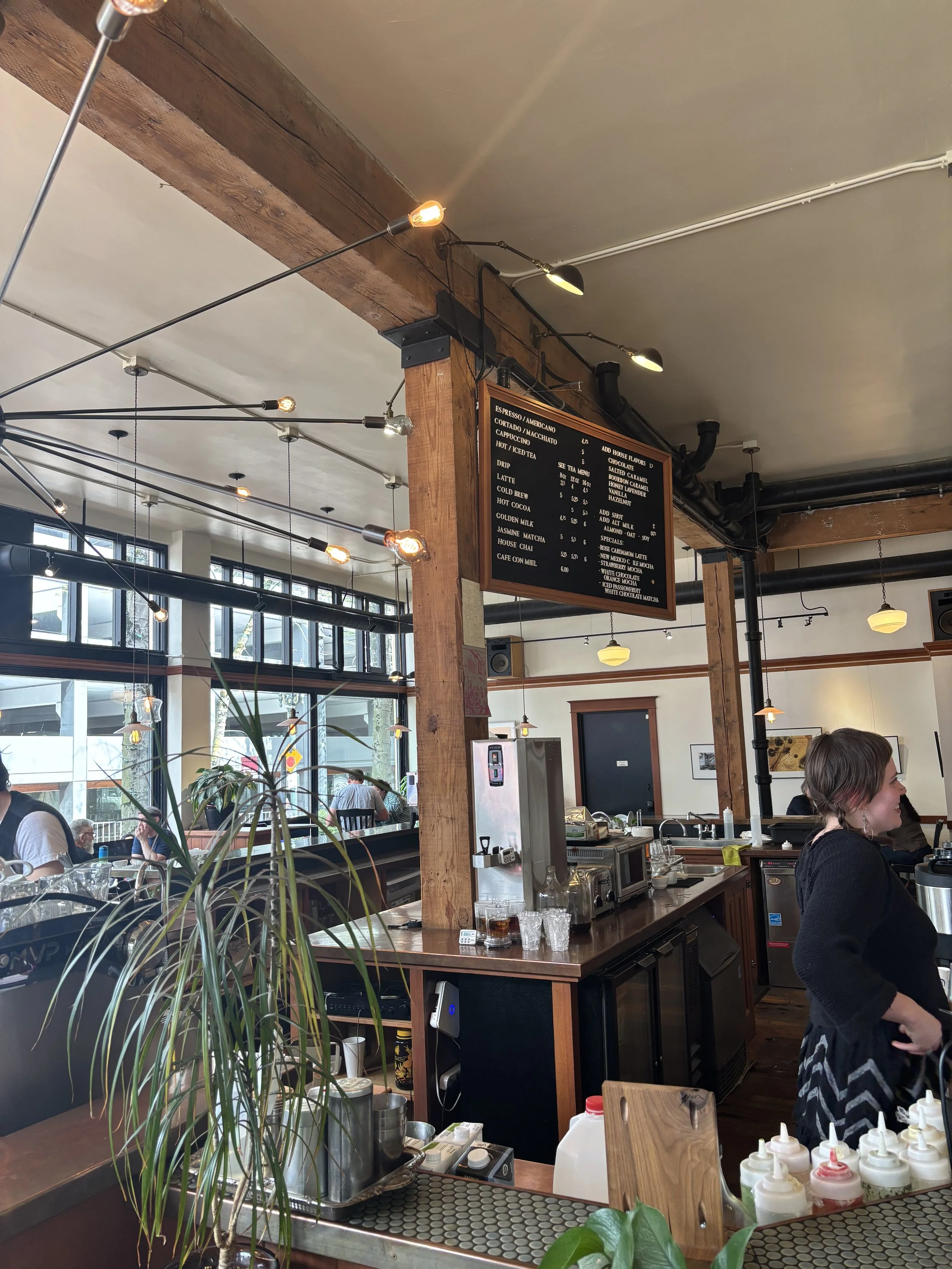 Interior of a cafe or restaurant with a wooden beam ceiling, large windows, and a black menu board showing coffee options. People are seated and a barista is working behind the counter. Bottles of sauces and drinks are visible on the counter.