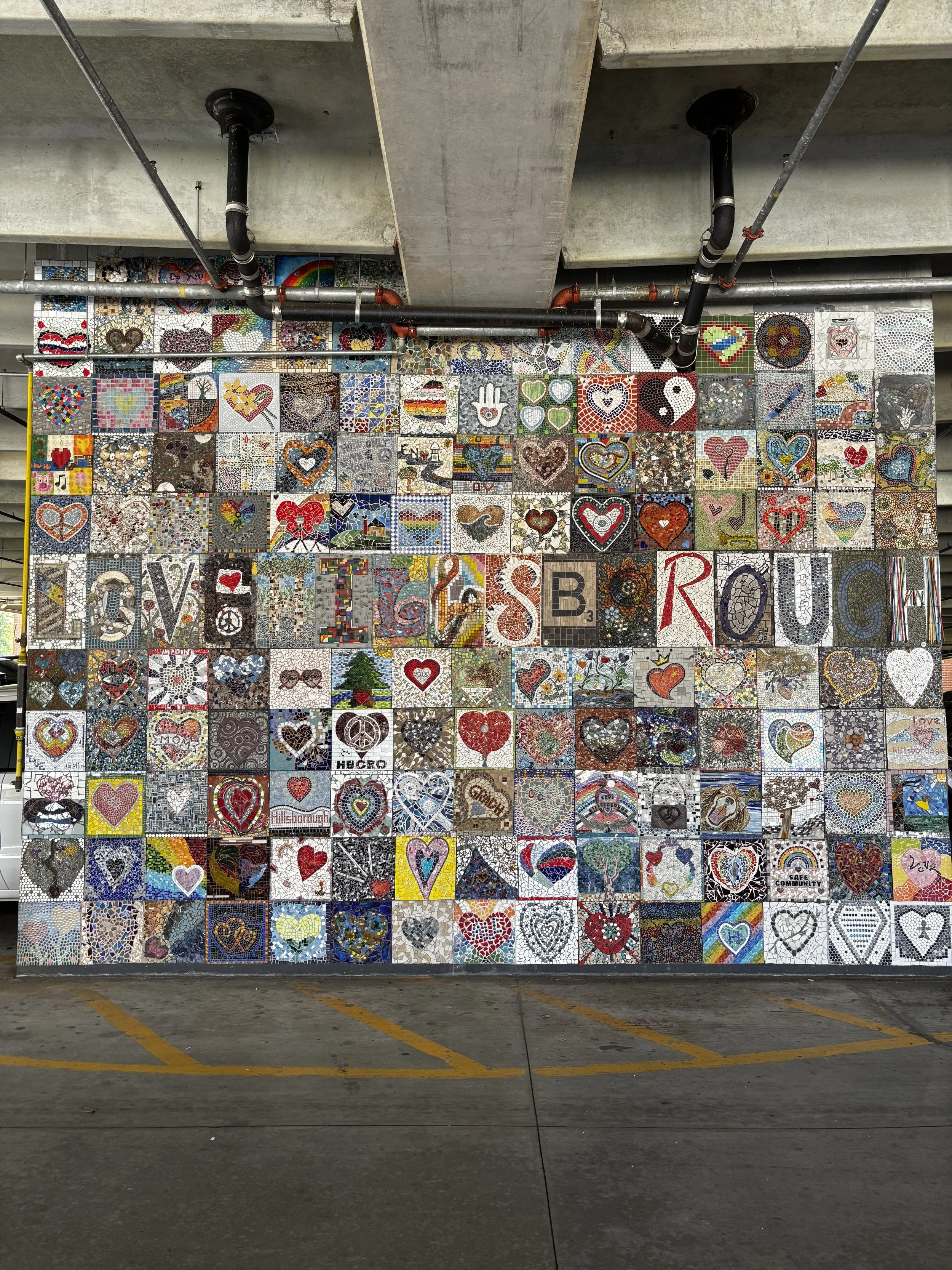 A large mosaic wall with numerous colorful heart designs, symbols, and images in a parking garage.