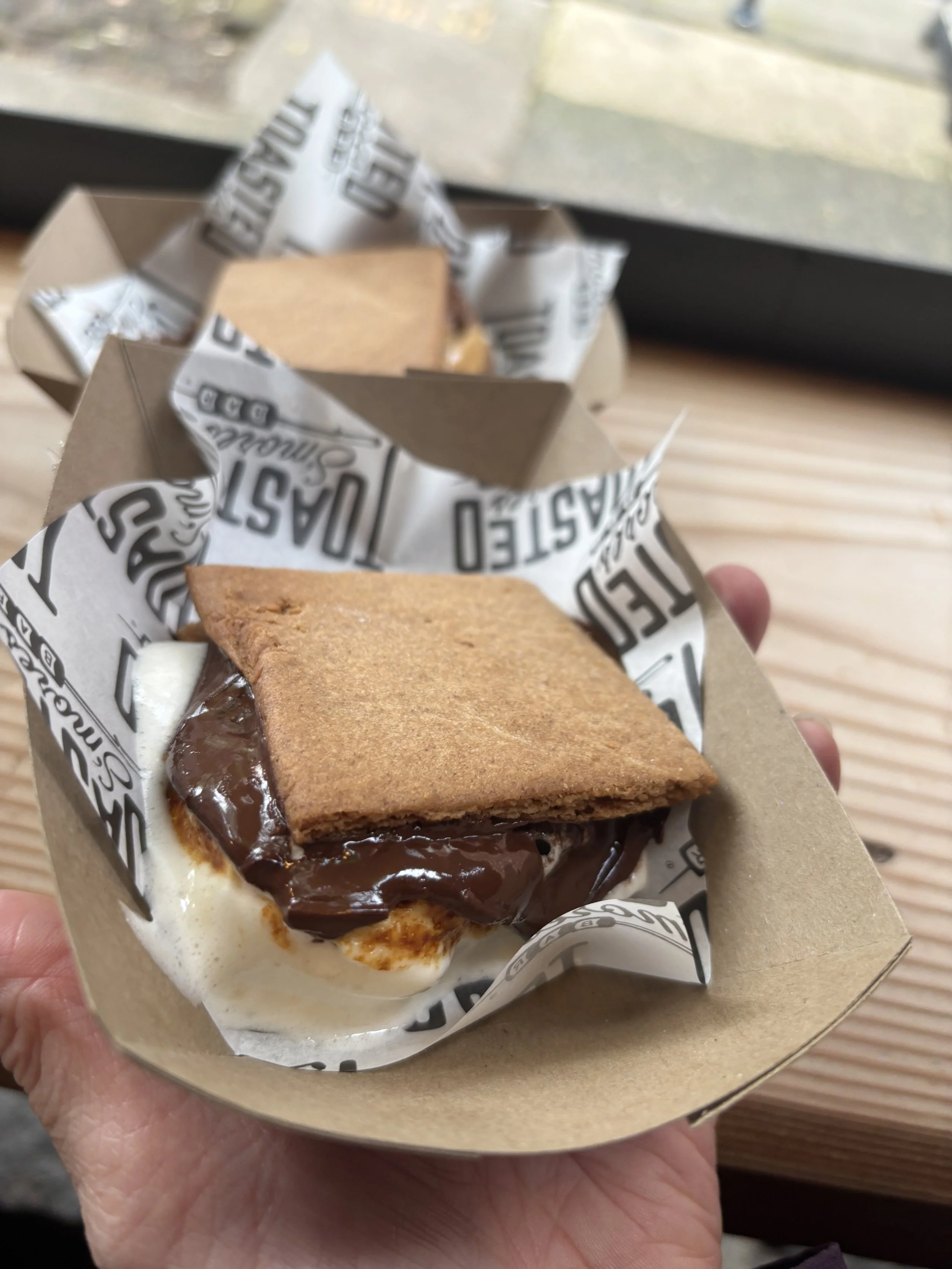 A hand holding a piece of cake with chocolate and vanilla layers inside, topped with a graham cracker cracker crust, in a paper wrapper with black and white text.