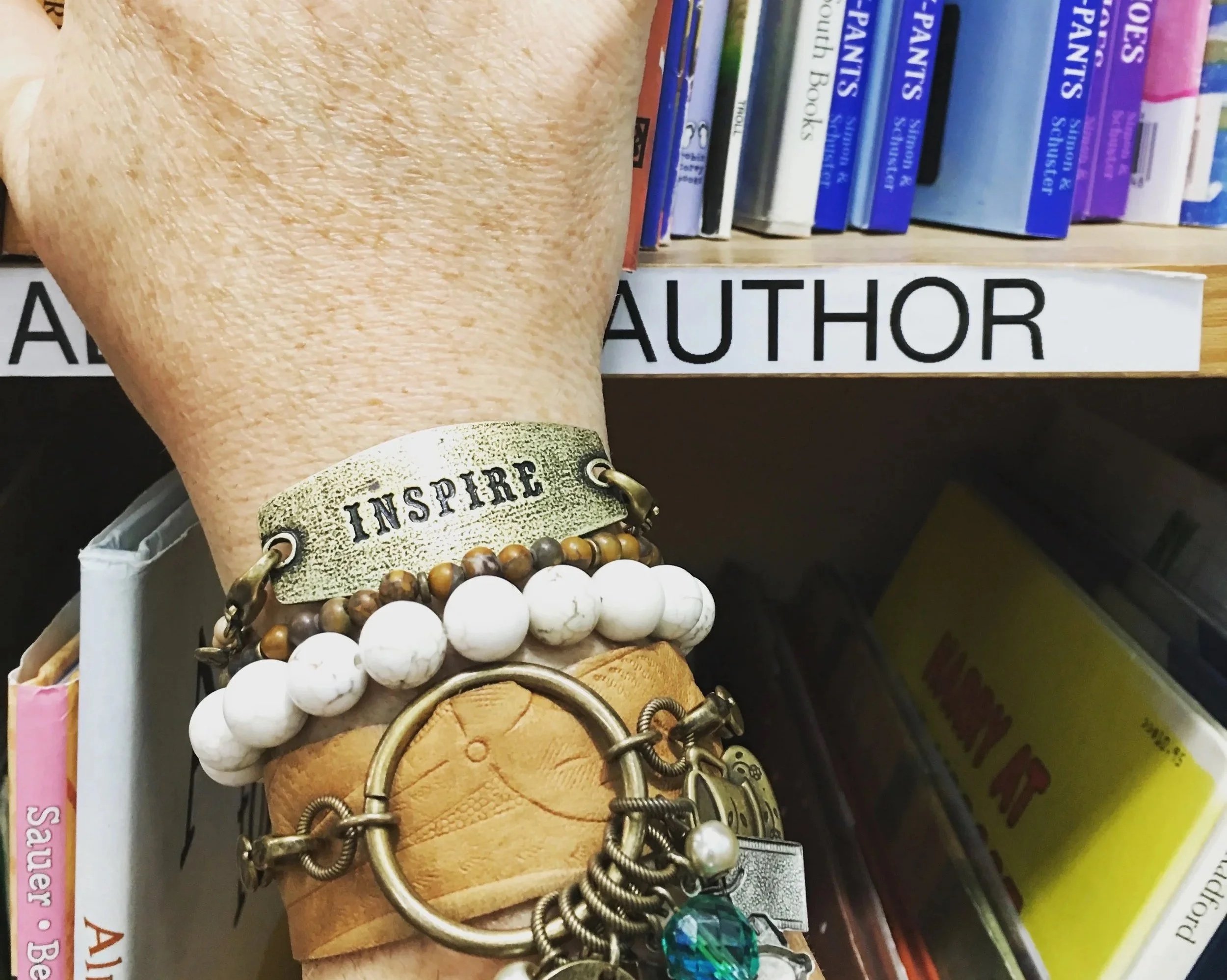 A shelf filled with multiple books, including titles related to health and personality. In front of the books, there is a collection of jewelry including a bracelet with the word 'INSPIRE' engraved on a gold-colored band, white and brown beaded bracelets, and a silver ring with a green stone.