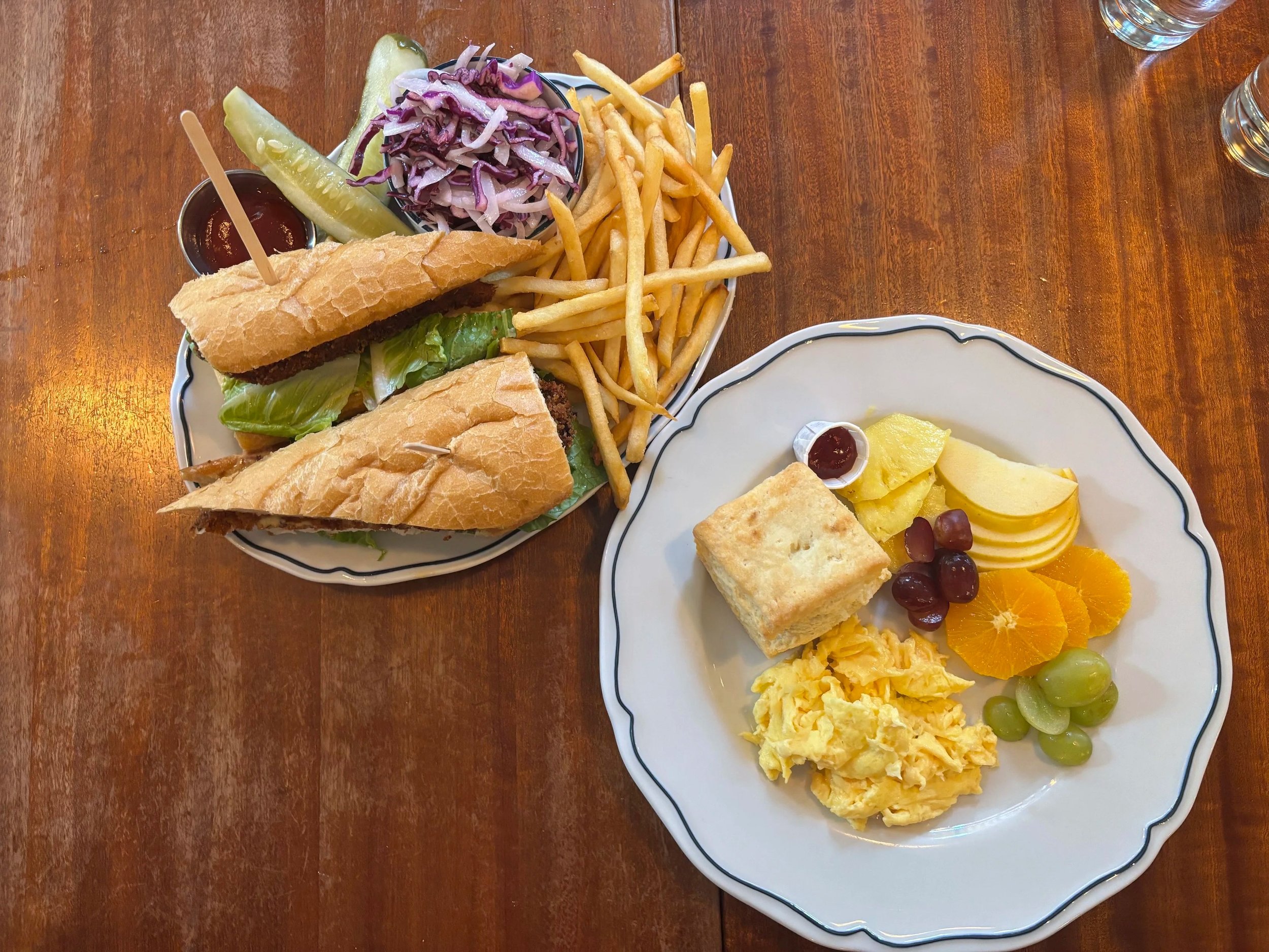 Plate of small sandwiches, French fries, coleslaw, and pickles with a small cup of barbecue sauce, alongside a plate of fruit salad with pineapple, grapes, orange slices, and a piece of cornbread.