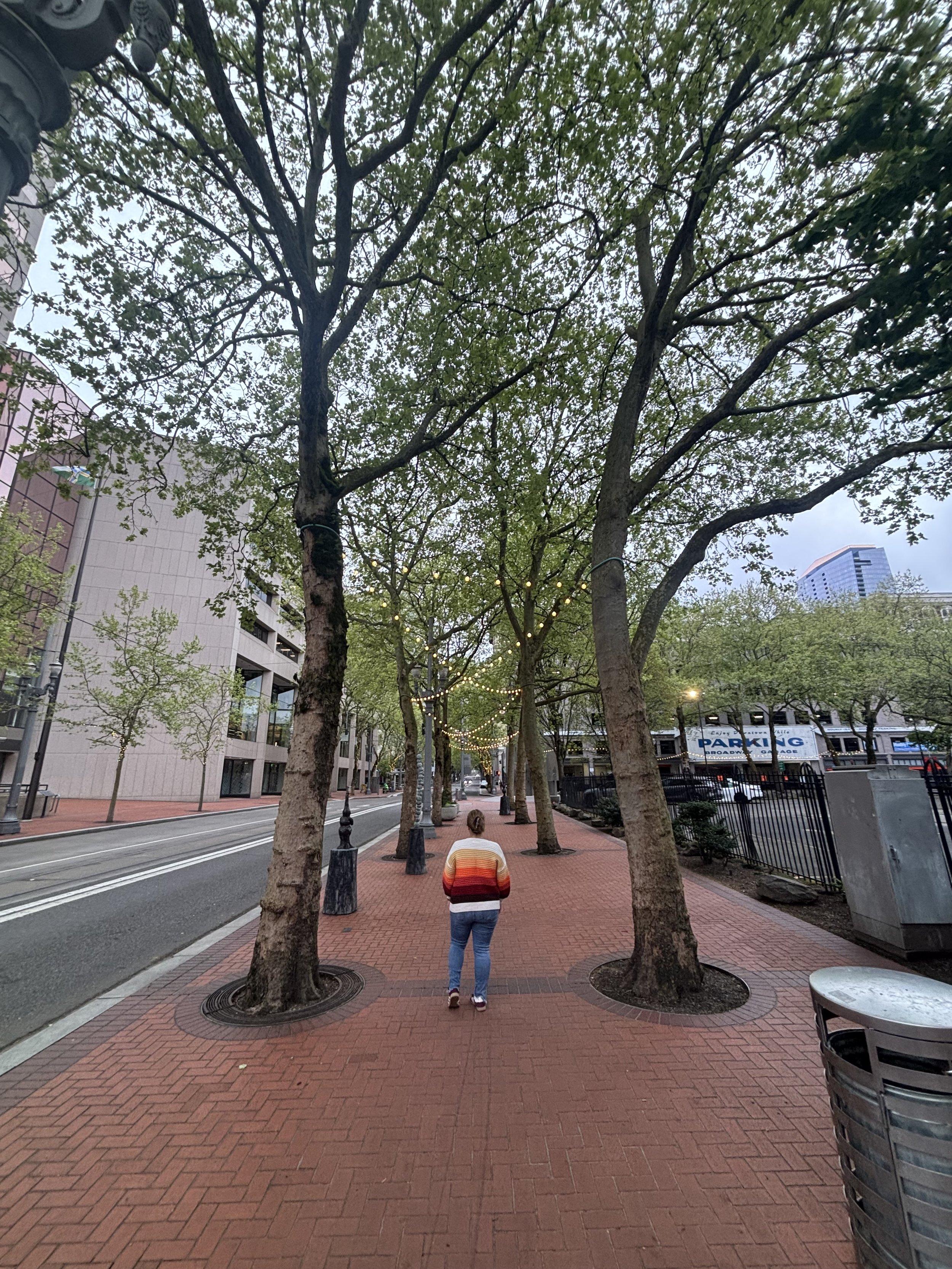 A person walking on a brick sidewalk lined with trees in an urban setting, with office buildings and a parking garage visible in the background.