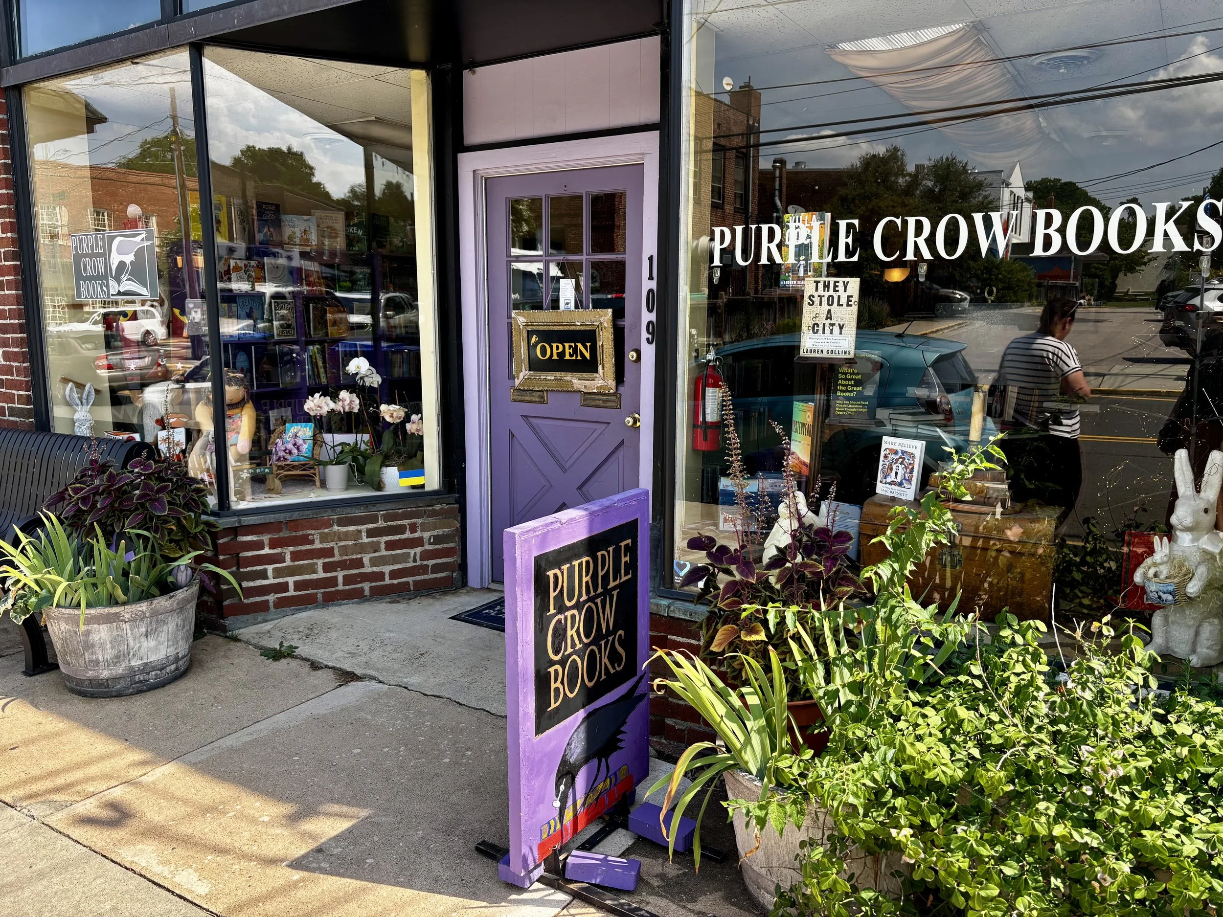 The storefront of Purple Crow Books with large glass windows, a purple door with a black border, and a purple sign with black letters. There are potted plants outside, and a chalkboard sign near the entrance that says "Purple Crow." Inside the window