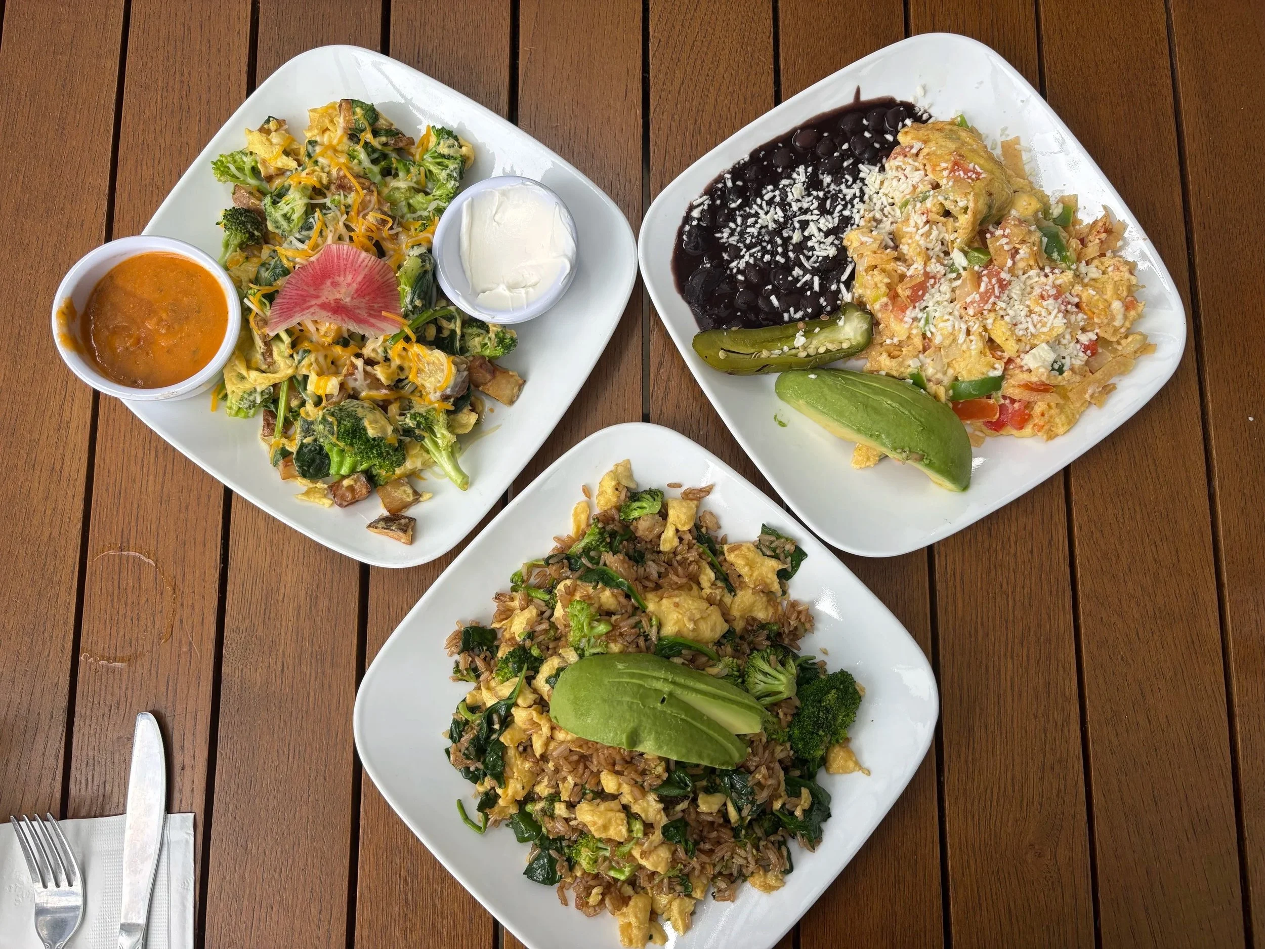 Three white rectangular plates on a wooden table with various dishes. The top left plate has broccoli, shredded cheese, and a dollop of sour cream. The top right plate has black beans with cheese, scrambled eggs with vegetables, and lime wedges. The bottom plate contains fried rice with vegetables and eggs, and lime wedges. There are forks and knives on napkins next to the plates.