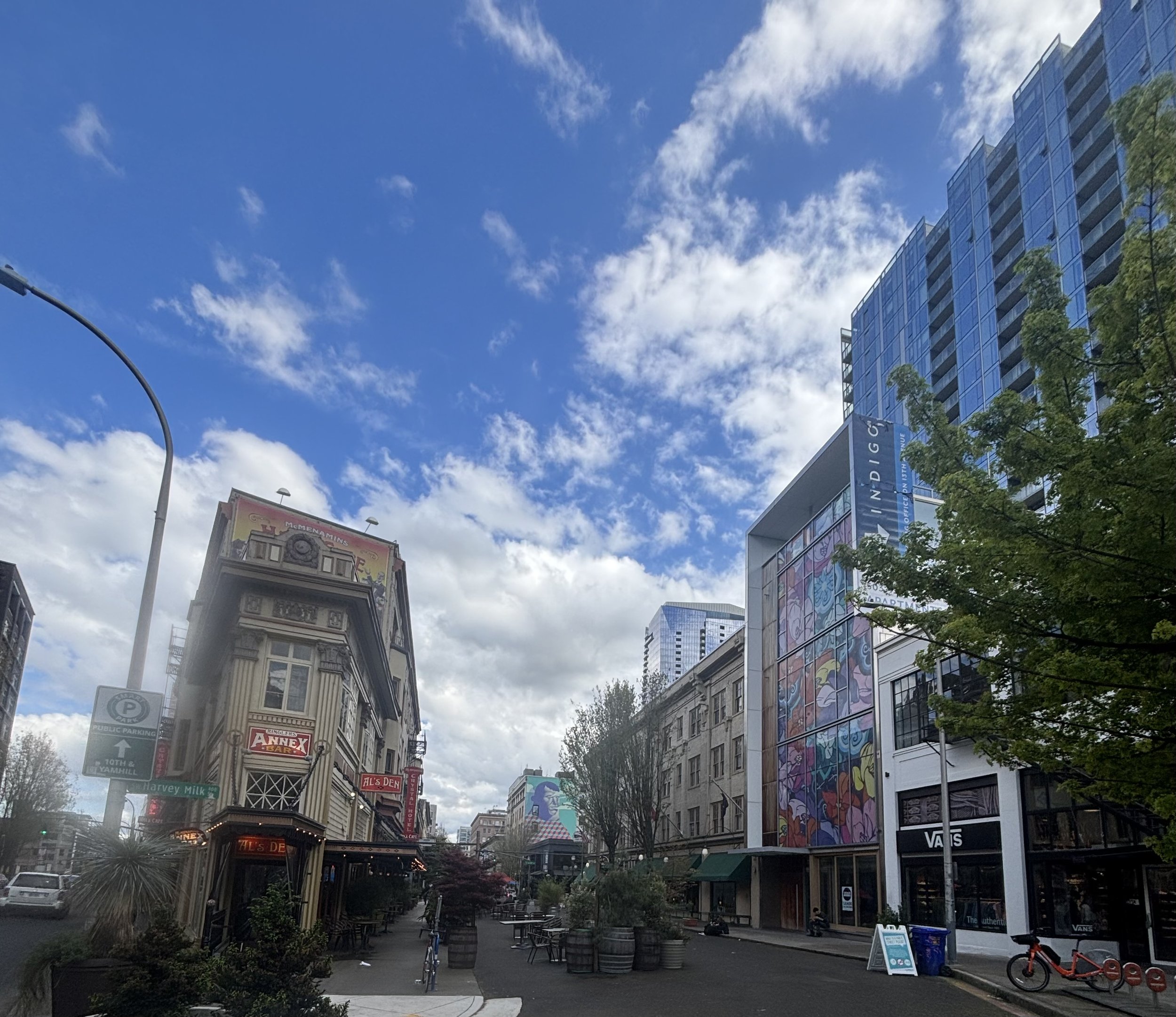 City street scene with tall modern glass buildings on the right, older architecture on the left, and a pedestrian area in the center. The sky is partly cloudy with patches of blue. There are signs for nearby establishments, streetlights, trees, outdo