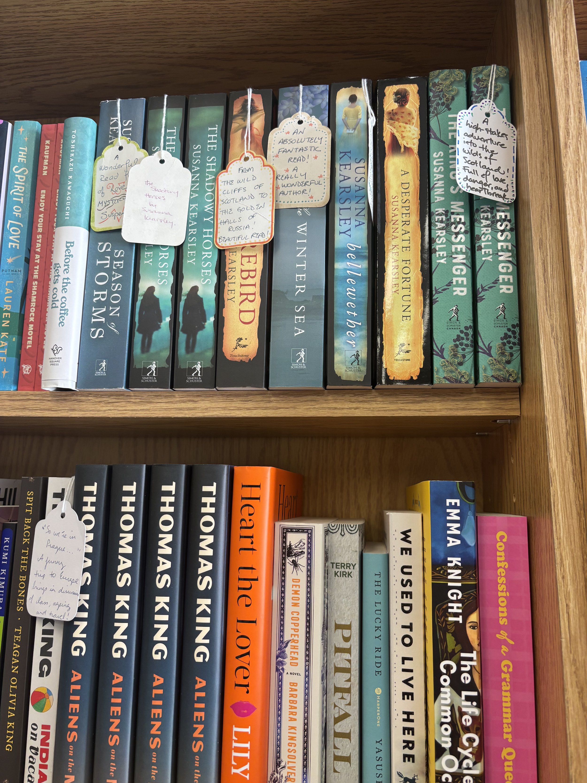 Shelf of books with various titles, including 'The Spirit of Love,' 'Before the Coffee Gets Cold,' 'The Shadow of the Sun,' 'Rebel,' 'A Desperate Fortune,' and others. Several of the books have handwritten tags attached, describing them as favorite reads, exciting adventures, or heartfelt stories.