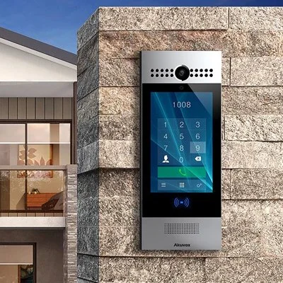 A modern intercom and security access panel mounted on an exterior brick wall of a building, featuring a screen, keypad, camera, and speaker.