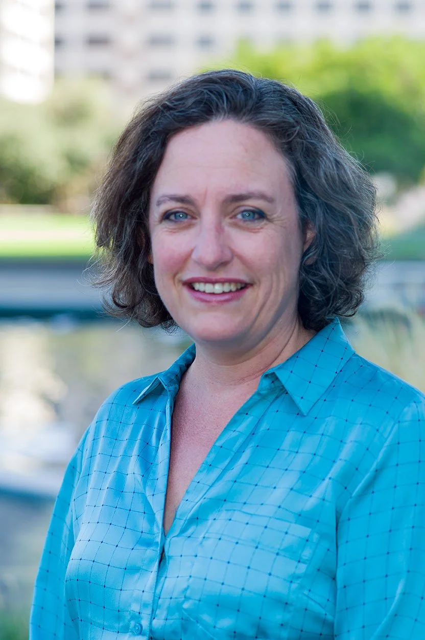 A woman with curly brown hair and blue eyes wearing a blue checkered shirt, standing outdoors with a blurred background of trees and buildings.
