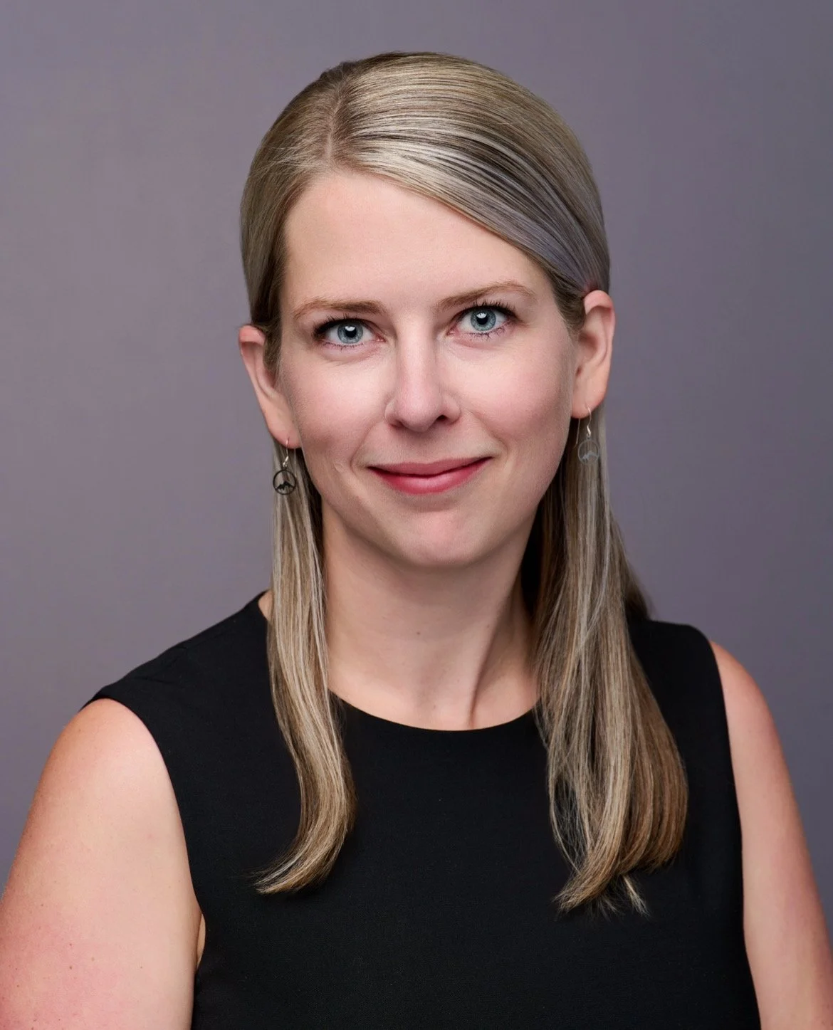 Headshot of a woman with blonde hair, blue eyes, wearing a black sleeveless top and earrings, against a gray background.