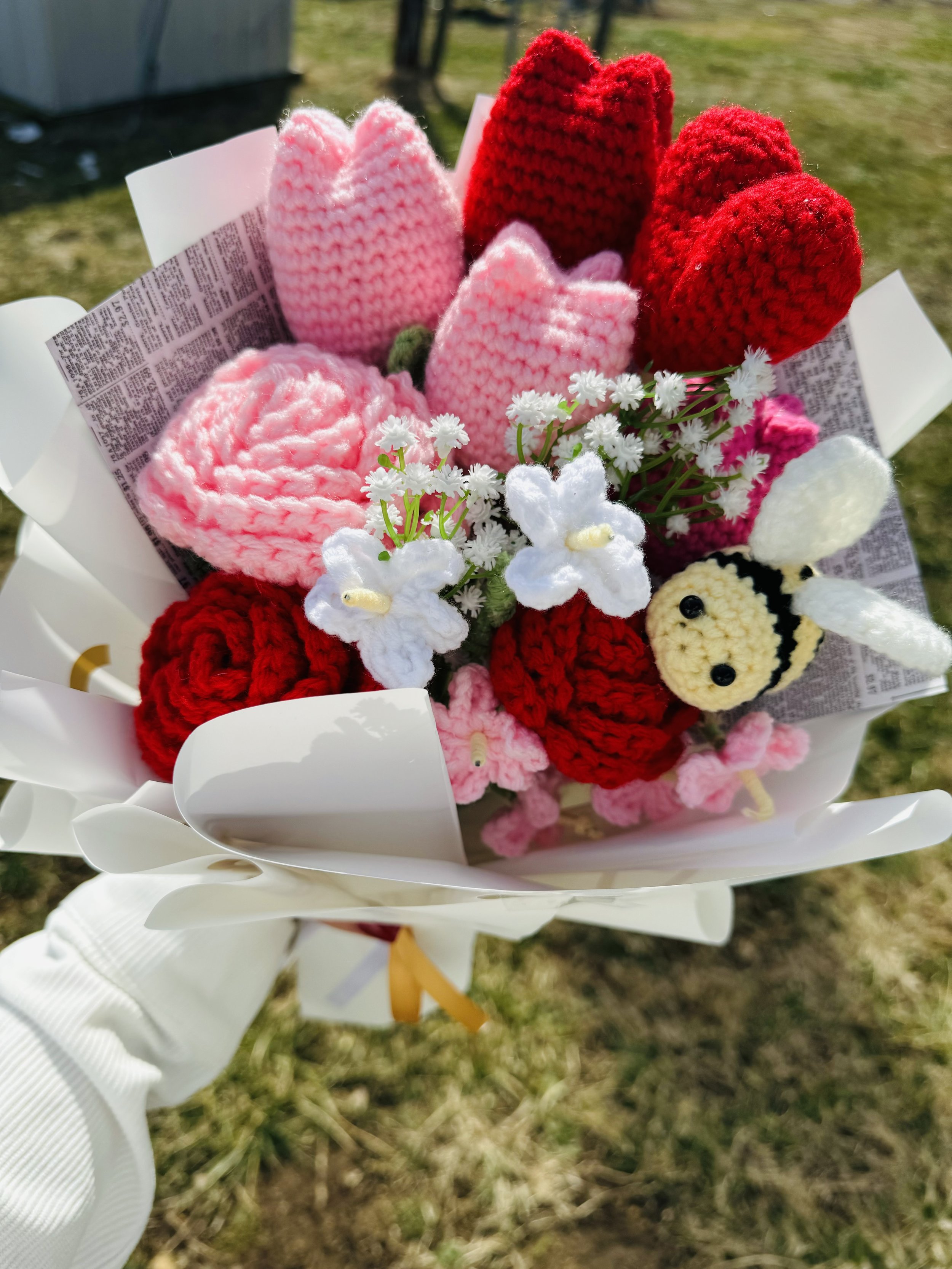 Close-up of a bouquet with crocheted flowers in pink, red, and white, along with white crocheted flowers resembling orchids, wrapped in white paper, outdoors on a grassy area.