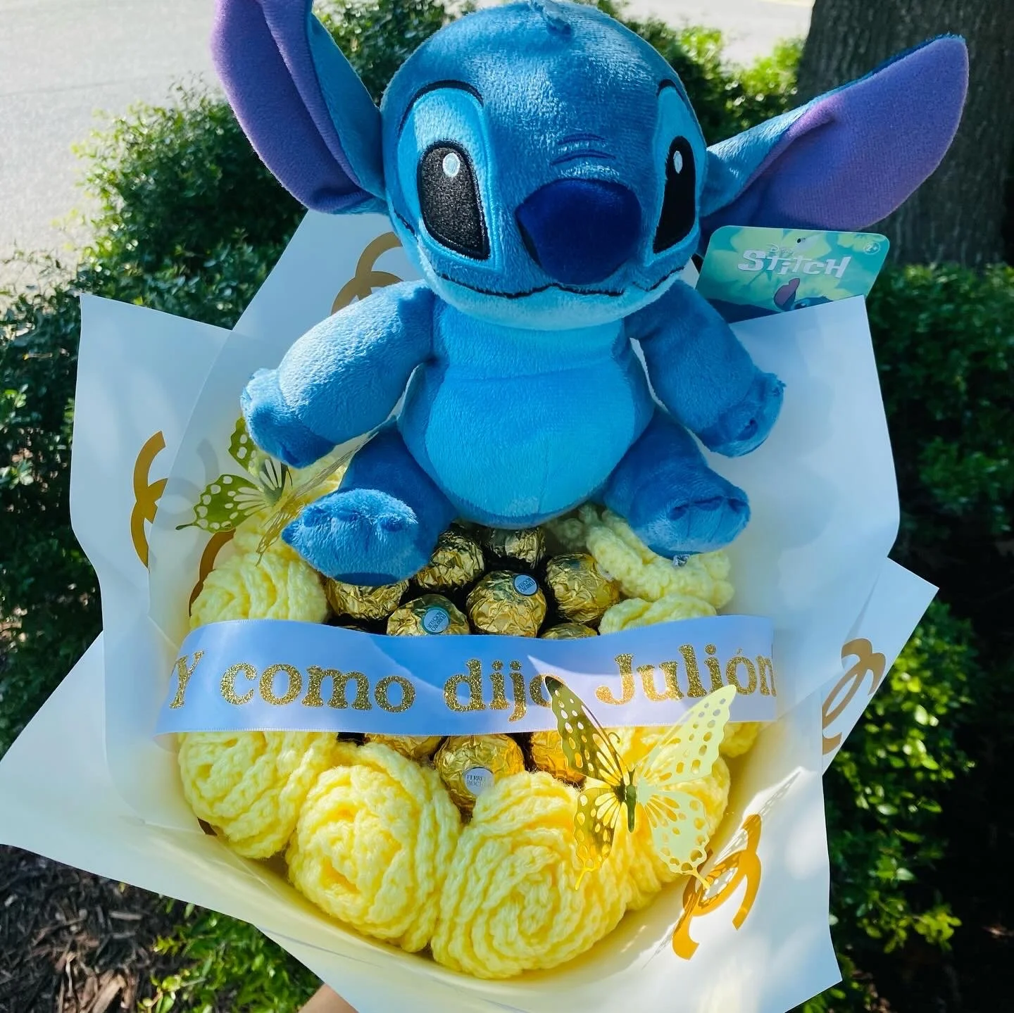A plush toy of Stitch from Disney's Lilo & Stitch sitting on a floral arrangement with chocolates wrapped in gold foil and yellow crocheted moss. There is a white banner with gold text that says, "Y como dijo Julión." The arrangement is decorated with yellow butterfly cutouts.
