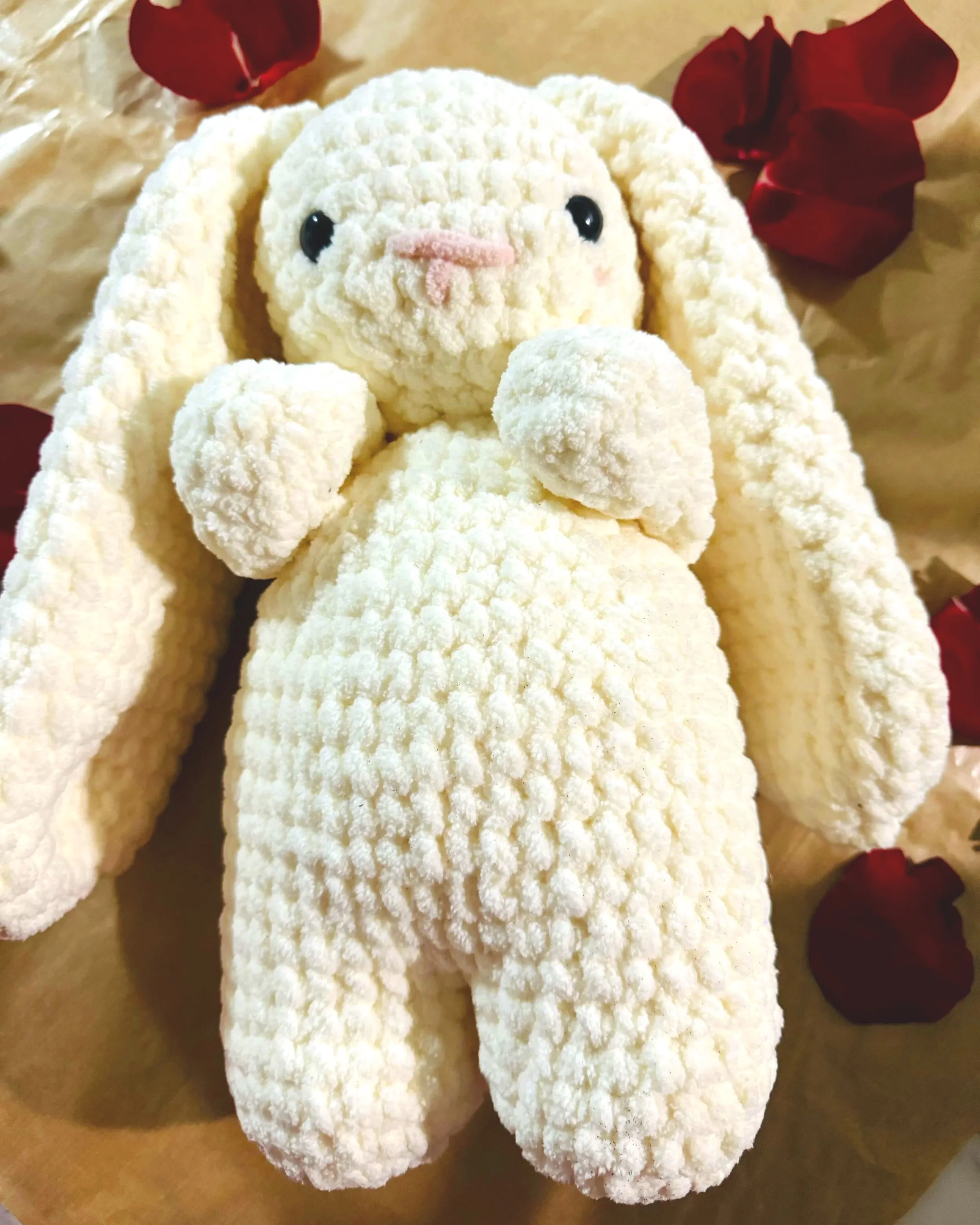 A plush stuffed animal resembling a white bunny with long ears, black eyes, and a pink nose, surrounded by red flower petals on a brown paper background.
