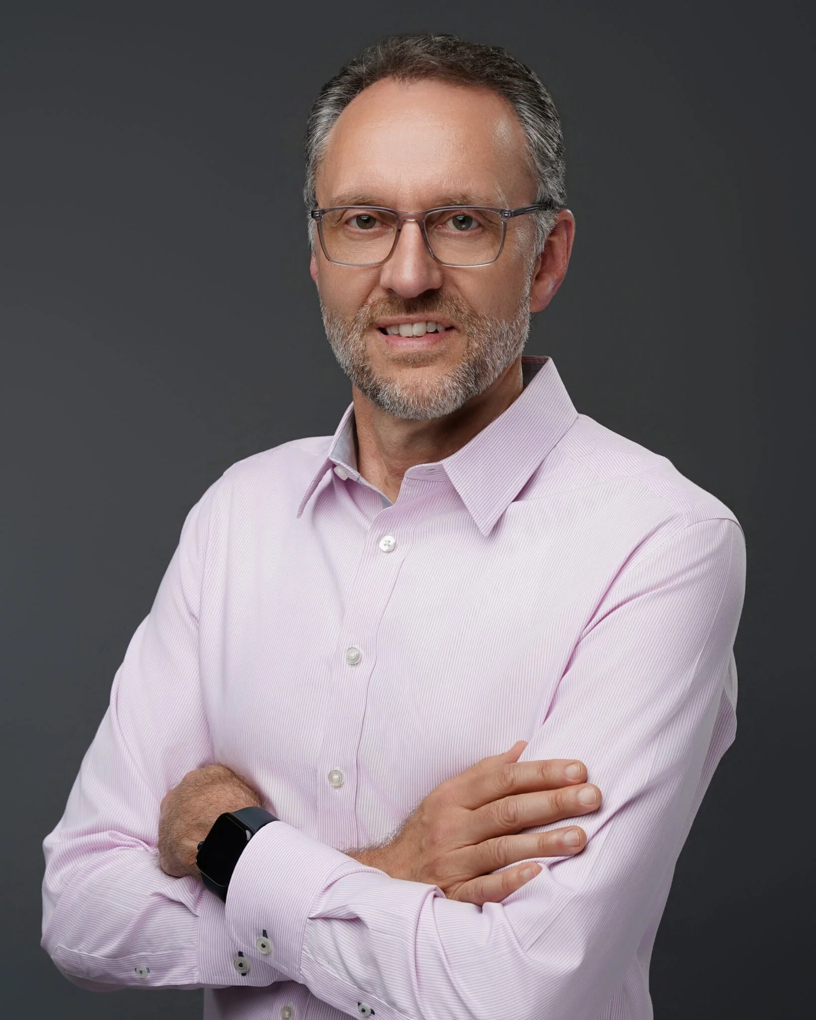 A middle-aged man with gray hair and beard wearing glasses and a light pink dress shirt, standing with arms crossed and smiling slightly against a dark gray background.