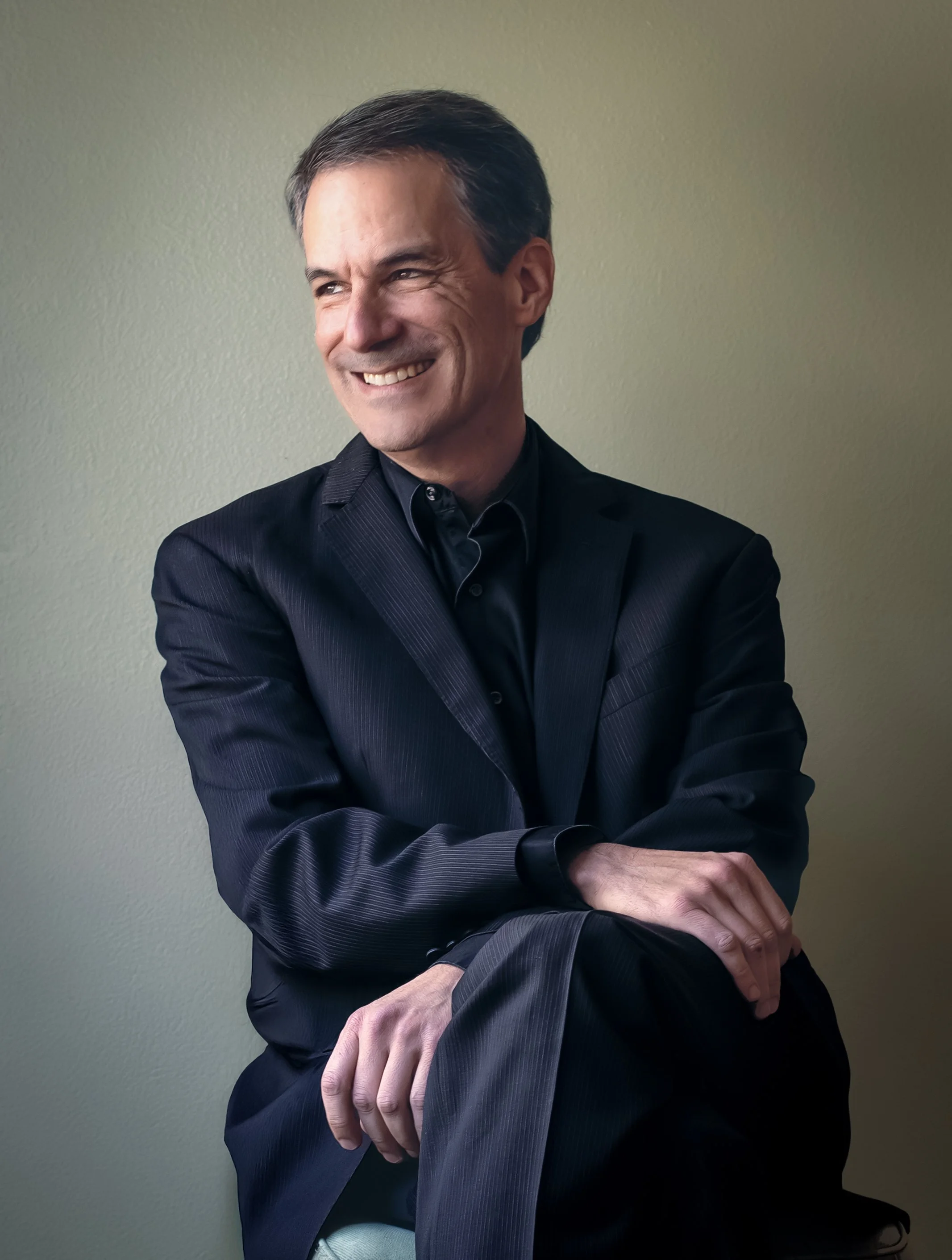 A man in a dark suit smiling and looking to his left with arms crossed, sitting against a plain beige wall.