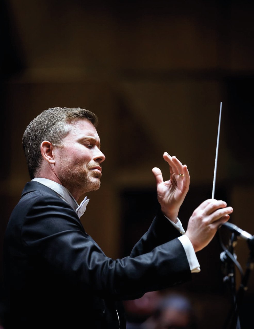 A male orchestra conductor wearing a tuxedo and bow tie, conducting an orchestra with a baton, with a serious expression.