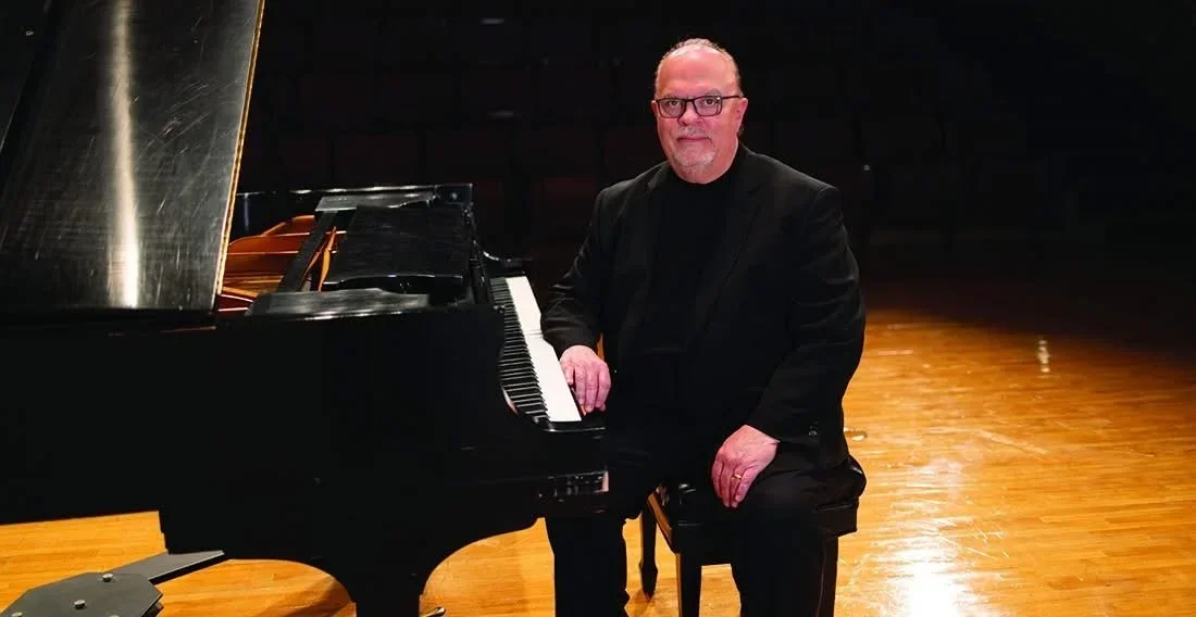 A man with glasses and a beard wearing a black suit and sitting at a grand piano on a wooden stage.