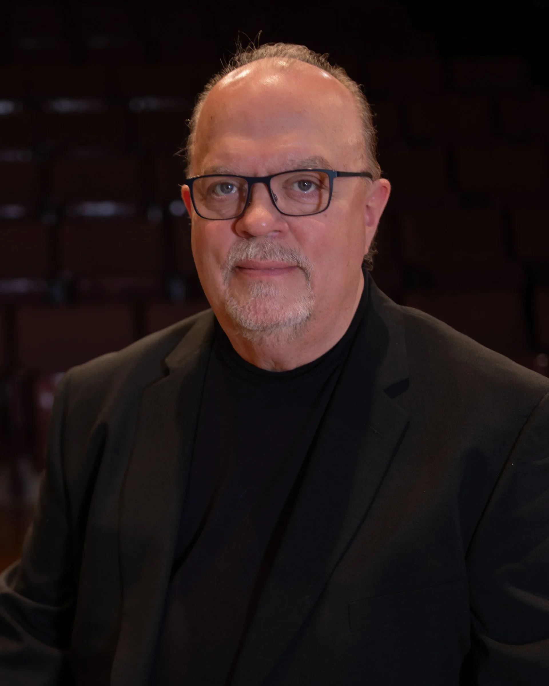 A close-up portrait of a middle-aged man with glasses, a light complexion, silver hair, and a beard, wearing a dark suit jacket, sitting indoors with a blurred background of dark seats.