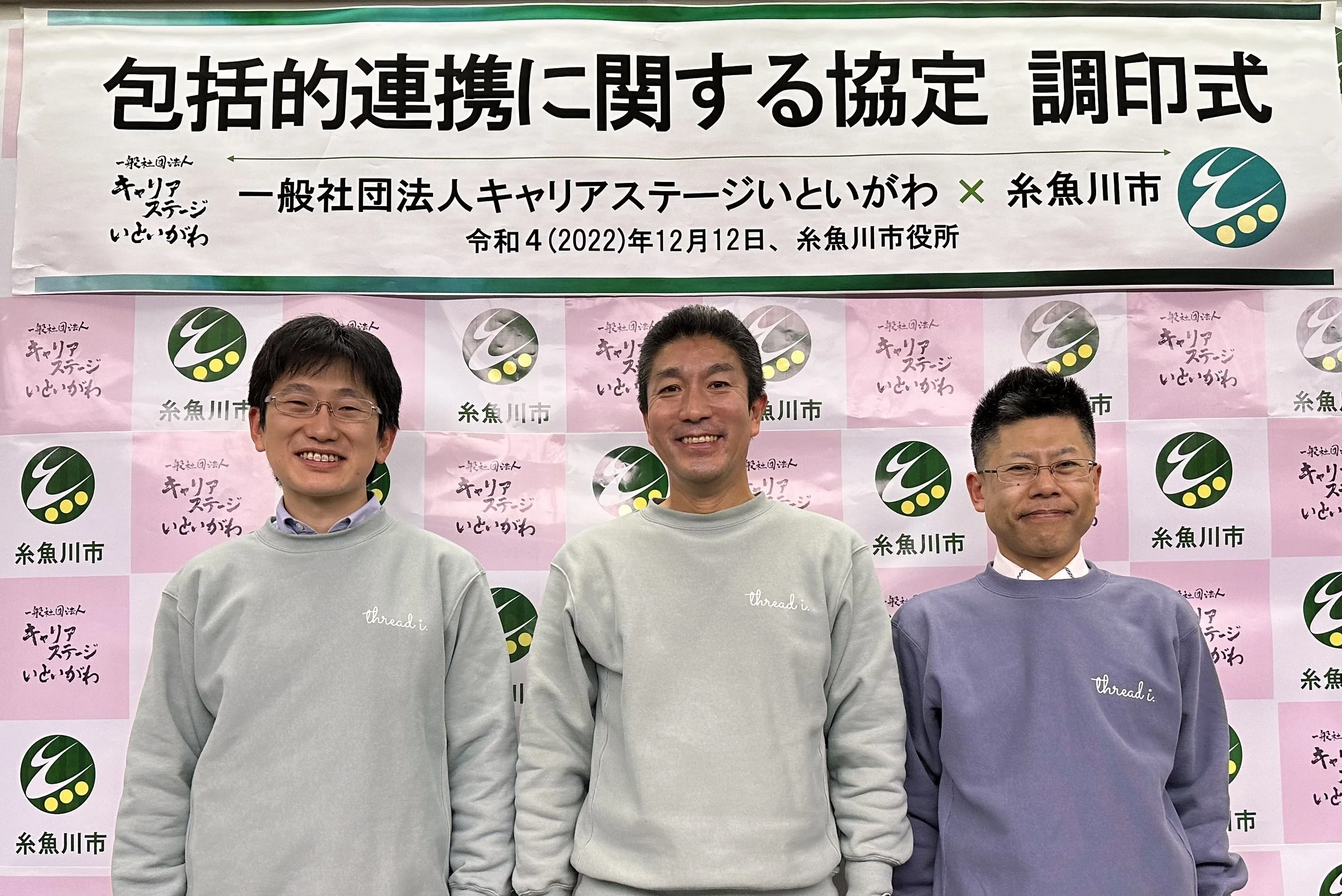 Three men standing in front of a backdrop with Japanese text and logos. They are smiling and wearing casual sweaters.