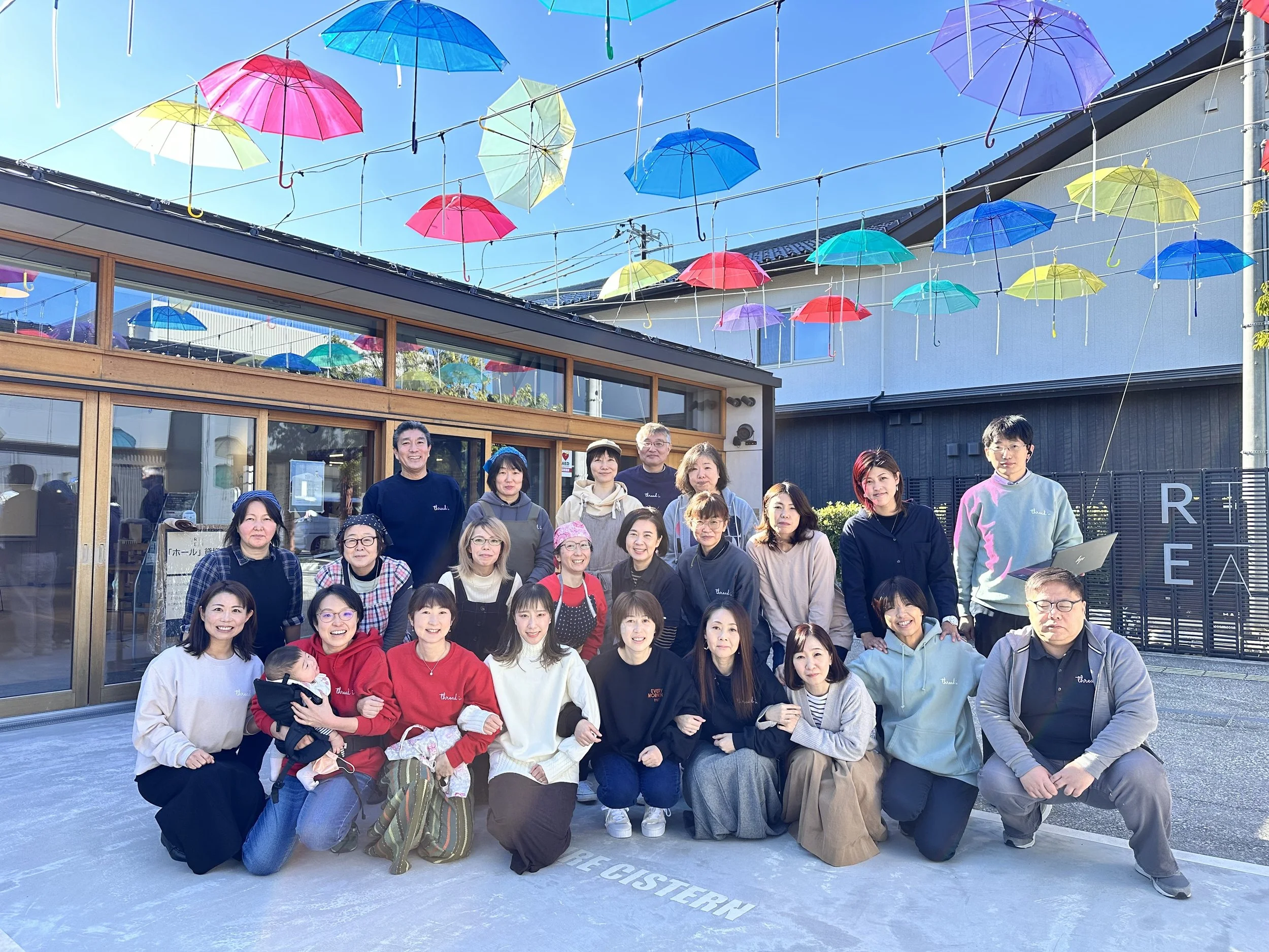 A group of people posing outside a building with colorful umbrellas hanging overhead against a bright blue sky.