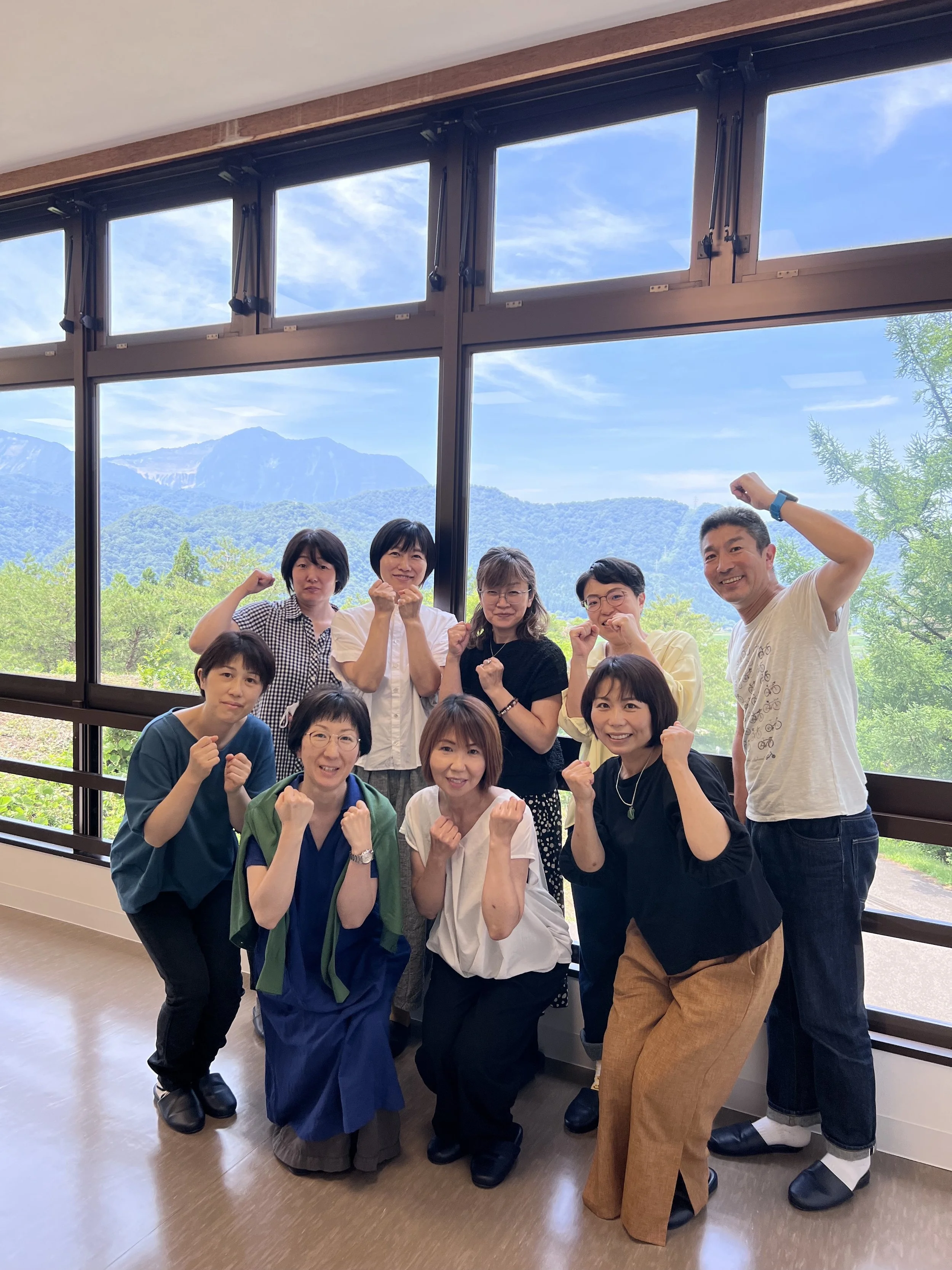 A group of nine people, mostly women and one man, standing inside a room with large windows showing mountains and greenery outside. They are posing with fists raised, smiling, and looking at the camera.