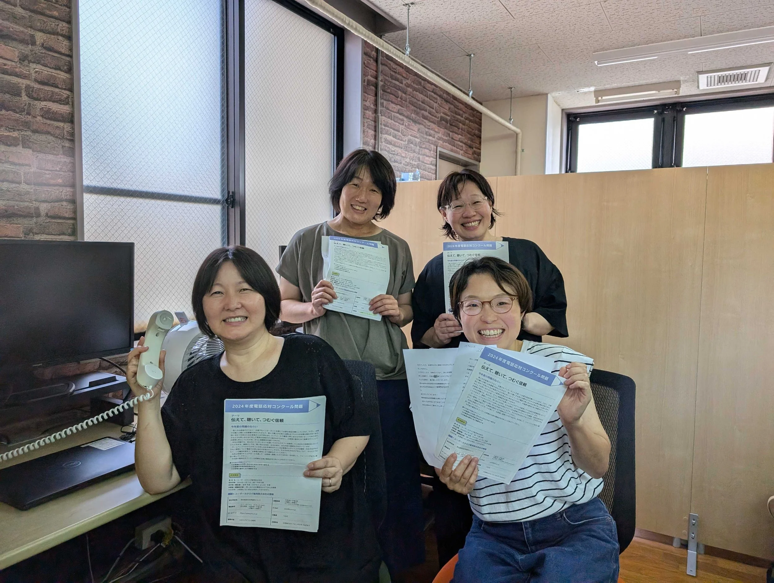A group of five smiling women in an office setting, holding papers and a telephone, with a brick wall and windows in the background.