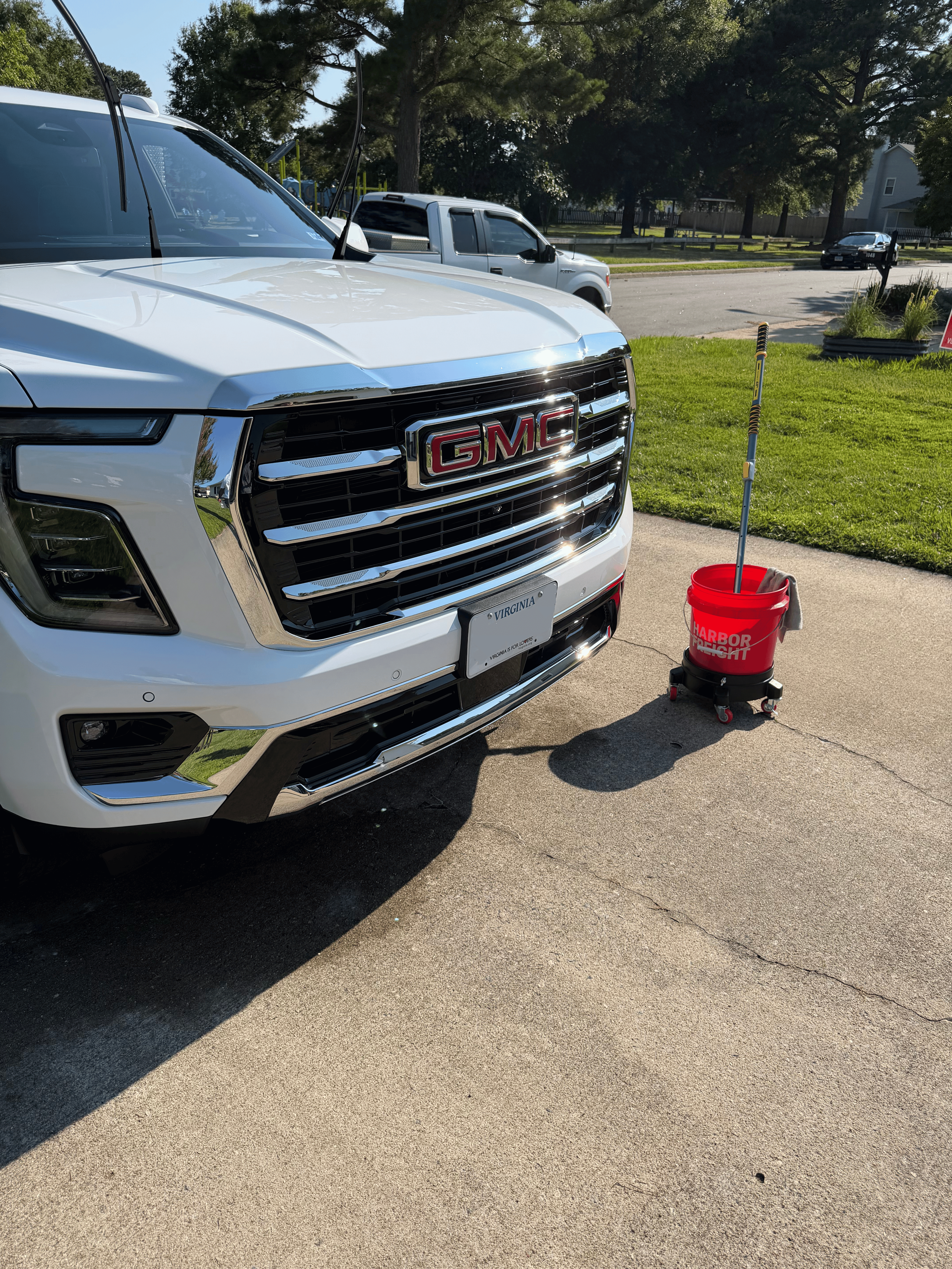White GMC vehicle parked on a street with a red Harbor Freight bucket next to it, and other cars in the background, trees, and houses.