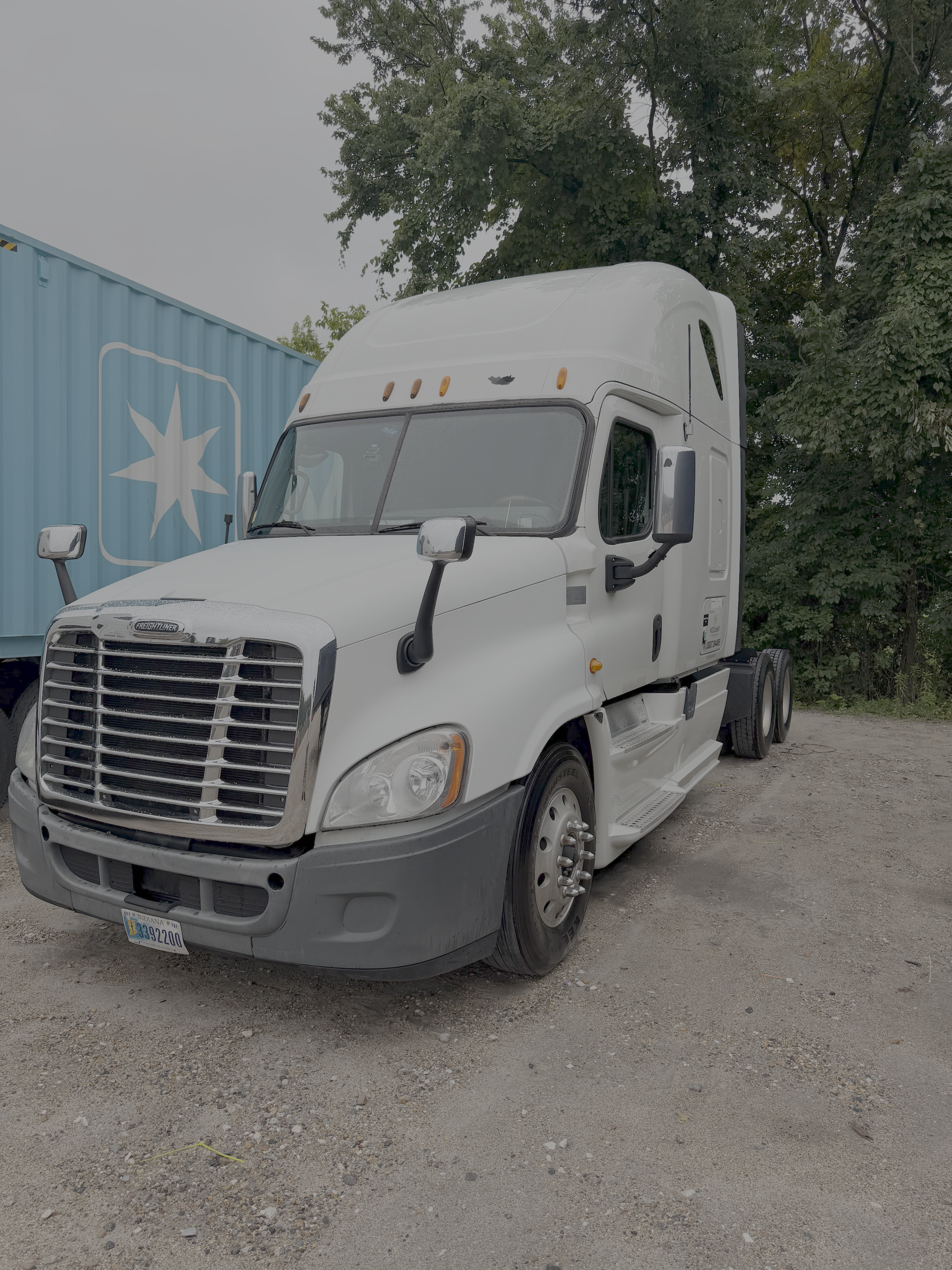 White semi-truck parked on a gravel surface with trees and a blue building in the background.