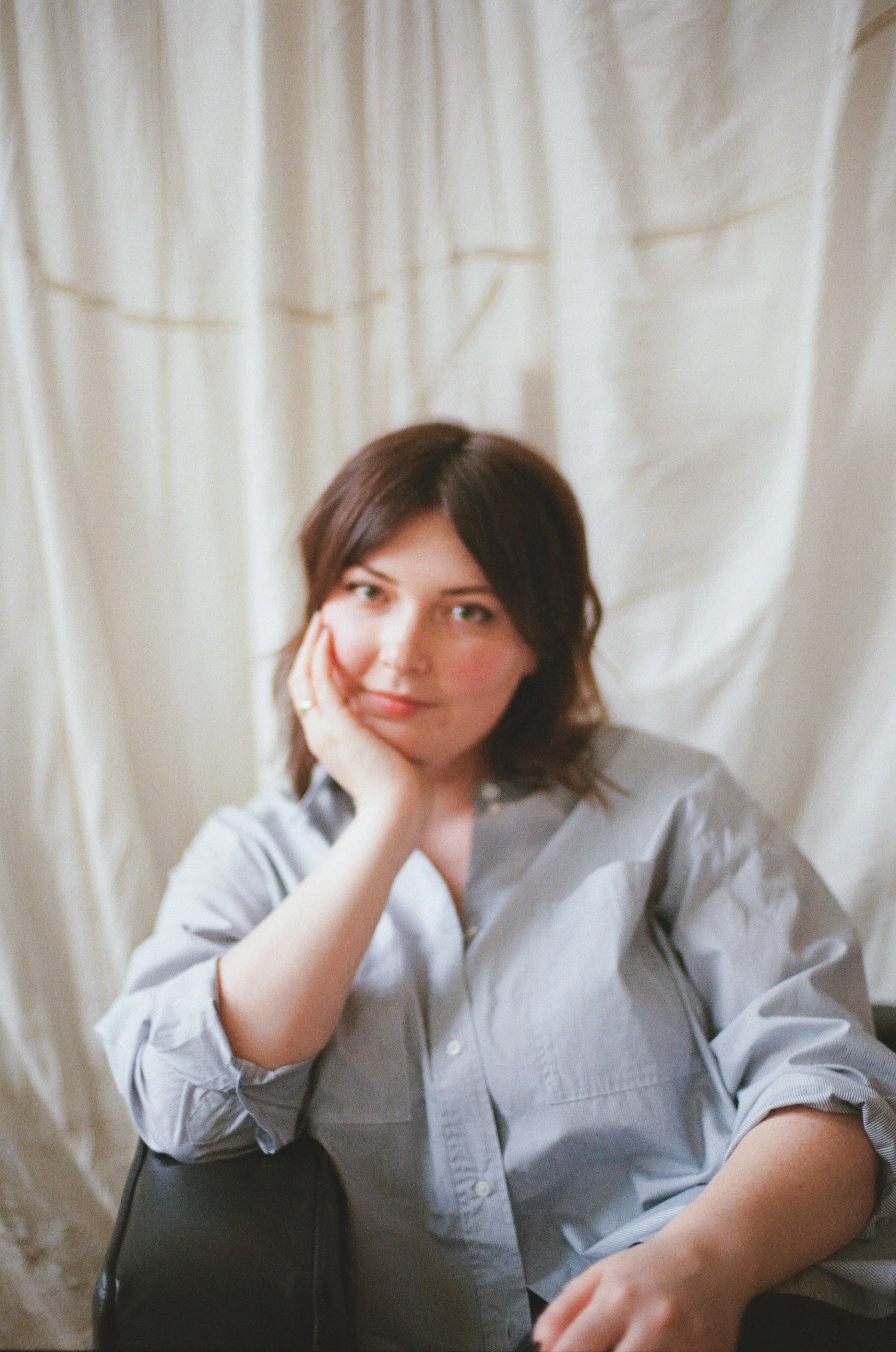 A woman with shoulder-length dark brown hair, wearing a light gray button-up shirt, seated on a black chair with her left elbow on the armrest and her head resting on her left hand, looking into the camera. The background features a cream-colored curtain.