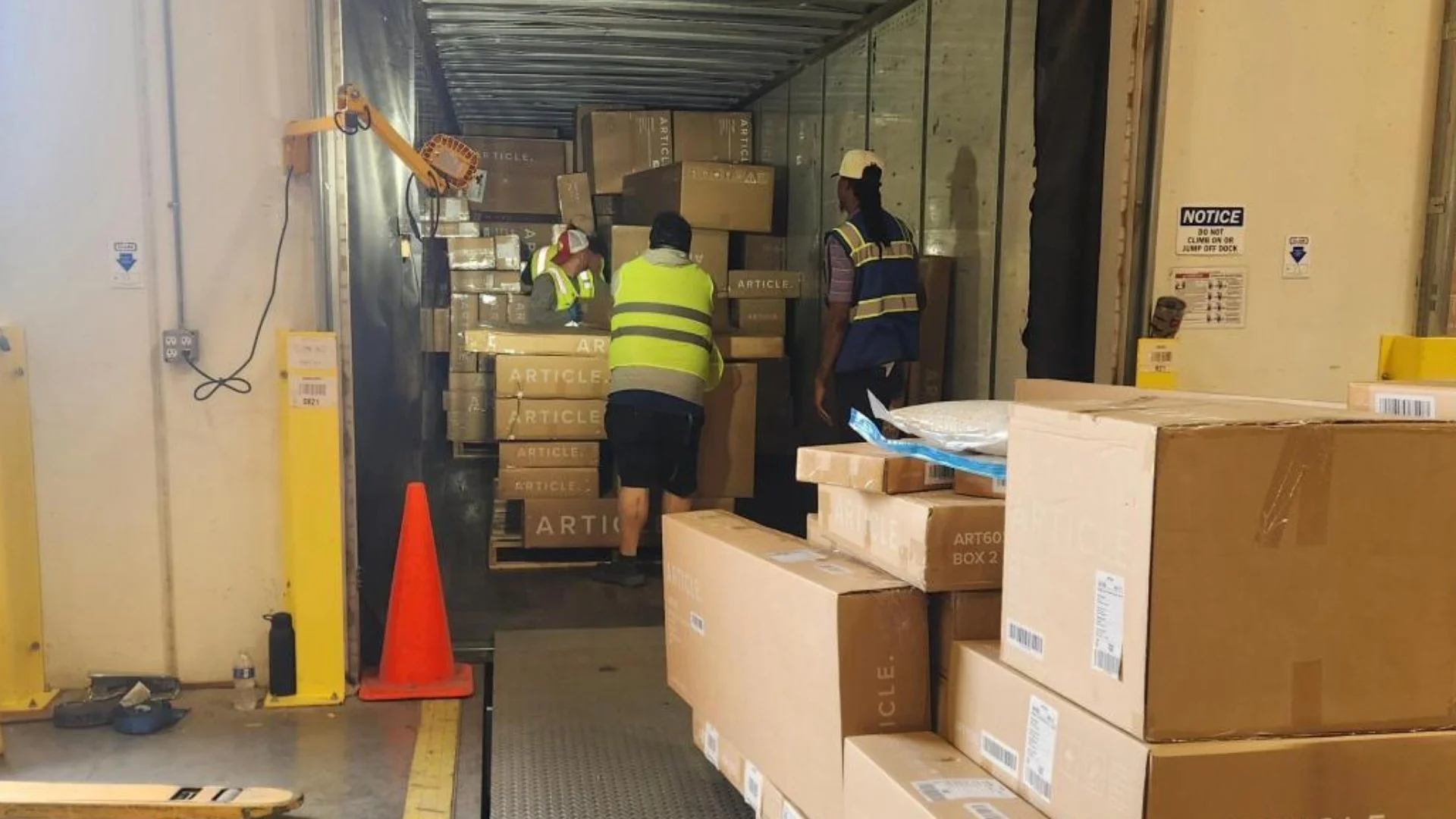 Warehouse workers in reflective vests load and organize cardboard boxes on a truck or storage area, with some boxes labeled 'ARTICLE' and others stacked on pallets.