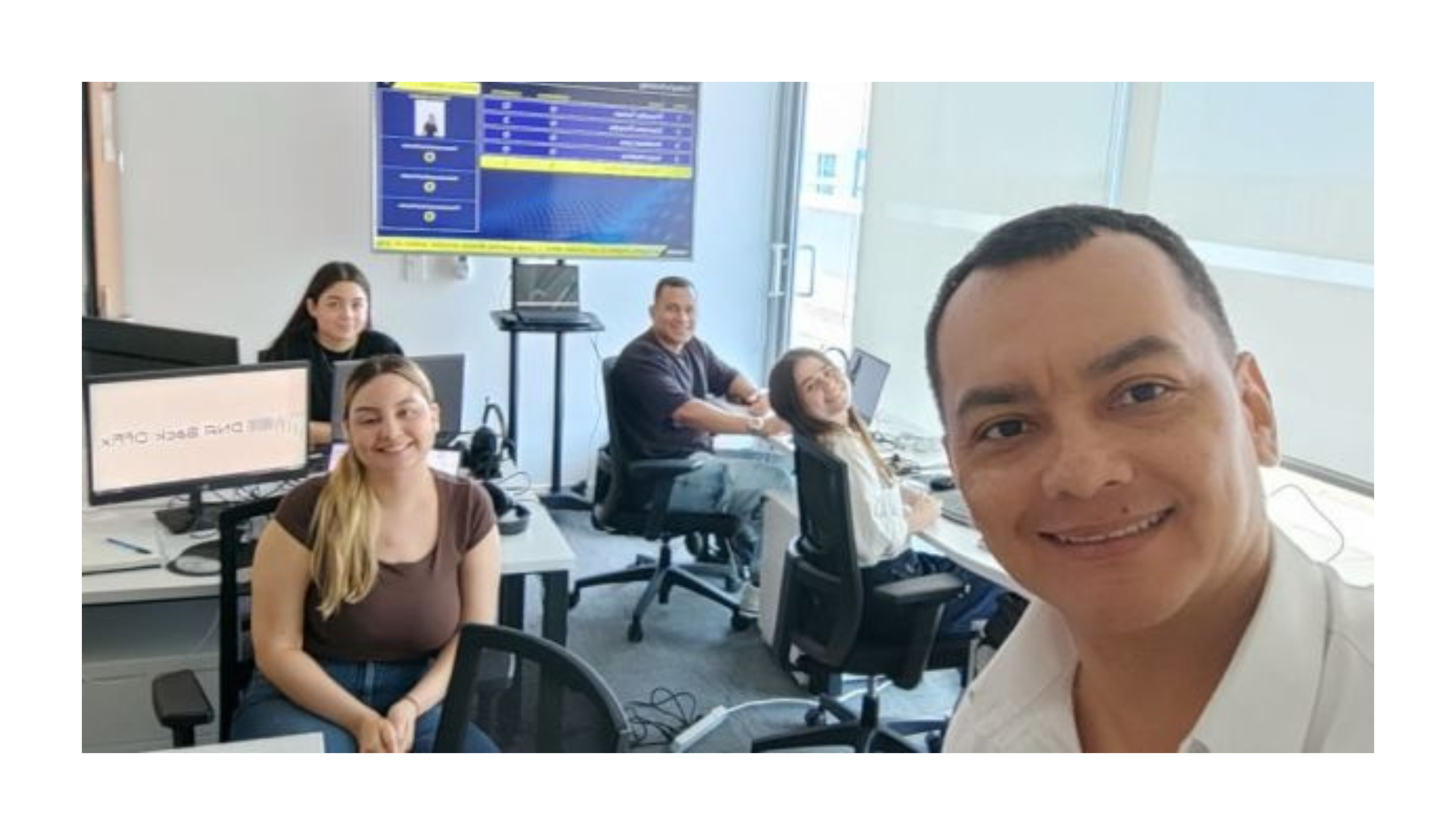 Group of five people sitting at desks in an office, smiling at the camera, with a large monitor displaying flight information in the background.