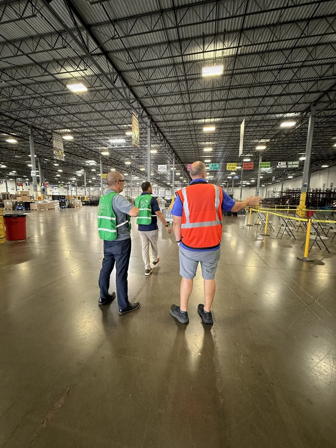 Three men walking inside a large warehouse or distribution center, two wearing green safety vests and one in an orange safety vest; there are shelves and signs in the background.