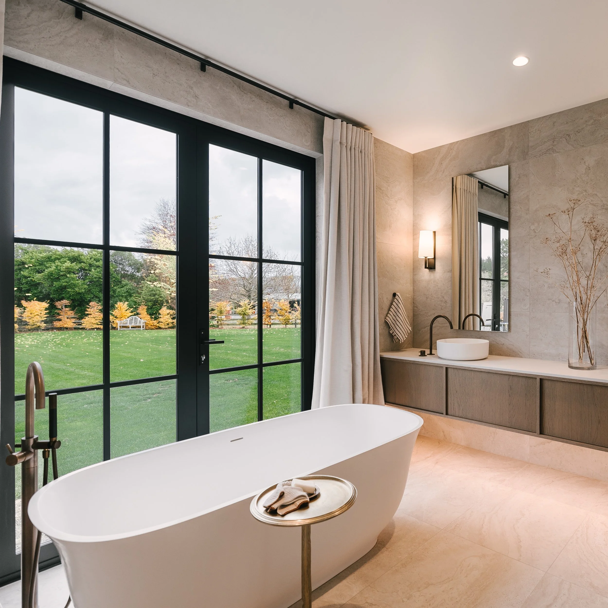 Modern bathroom with a freestanding white bathtub, double sink vanity with a mirror, beige tiled walls, large windows, and a view of green trees outside.