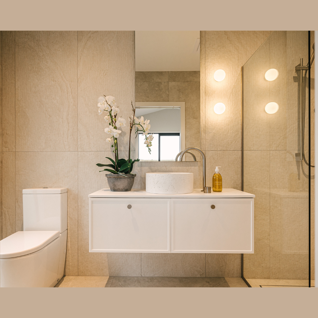 Modern bathroom with beige tiled walls, white floating vanity with vessel sink, orchid plant, soap dispenser, mirror, and shower with glass door.