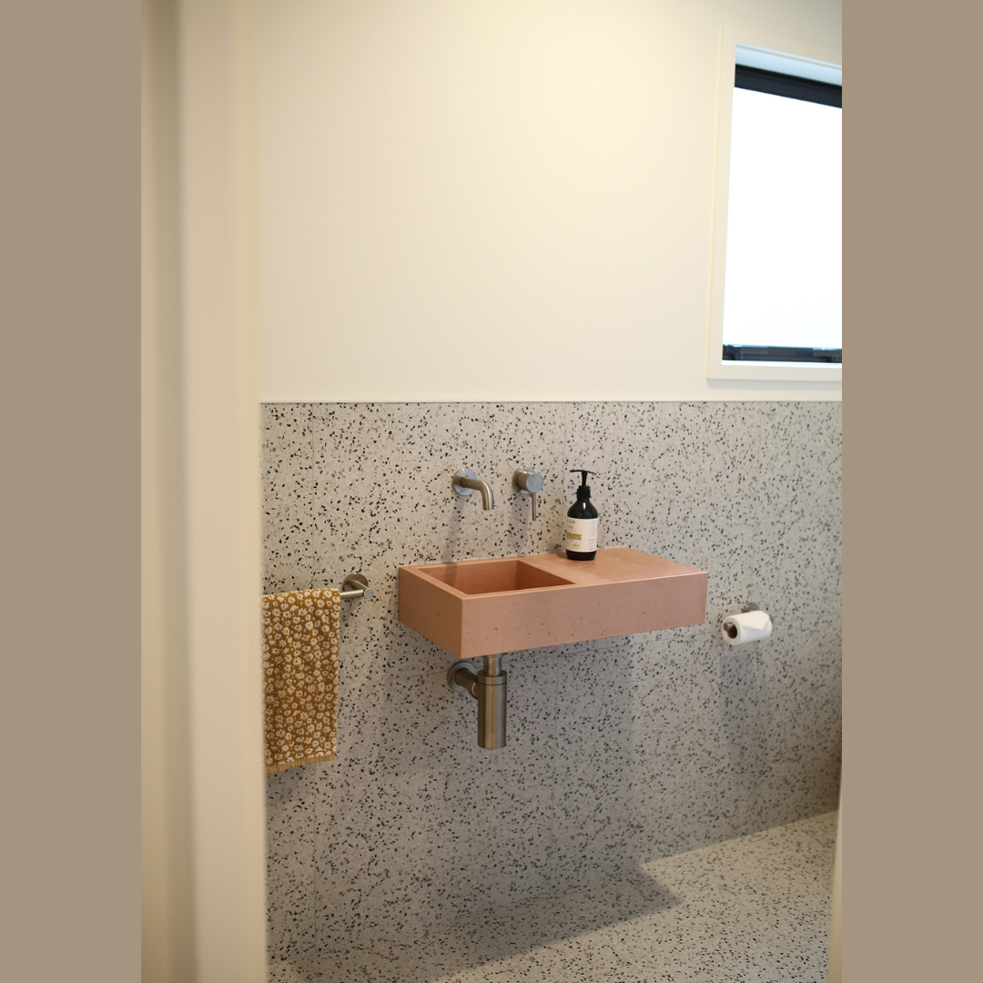 A small, modern bathroom with a pink rectangular sink mounted on a speckled gray wall, a black soap dispenser on the sink, a yellow patterned towel hanging on a wall-mounted towel holder, a toilet paper roll nearby, and a window providing natural lig