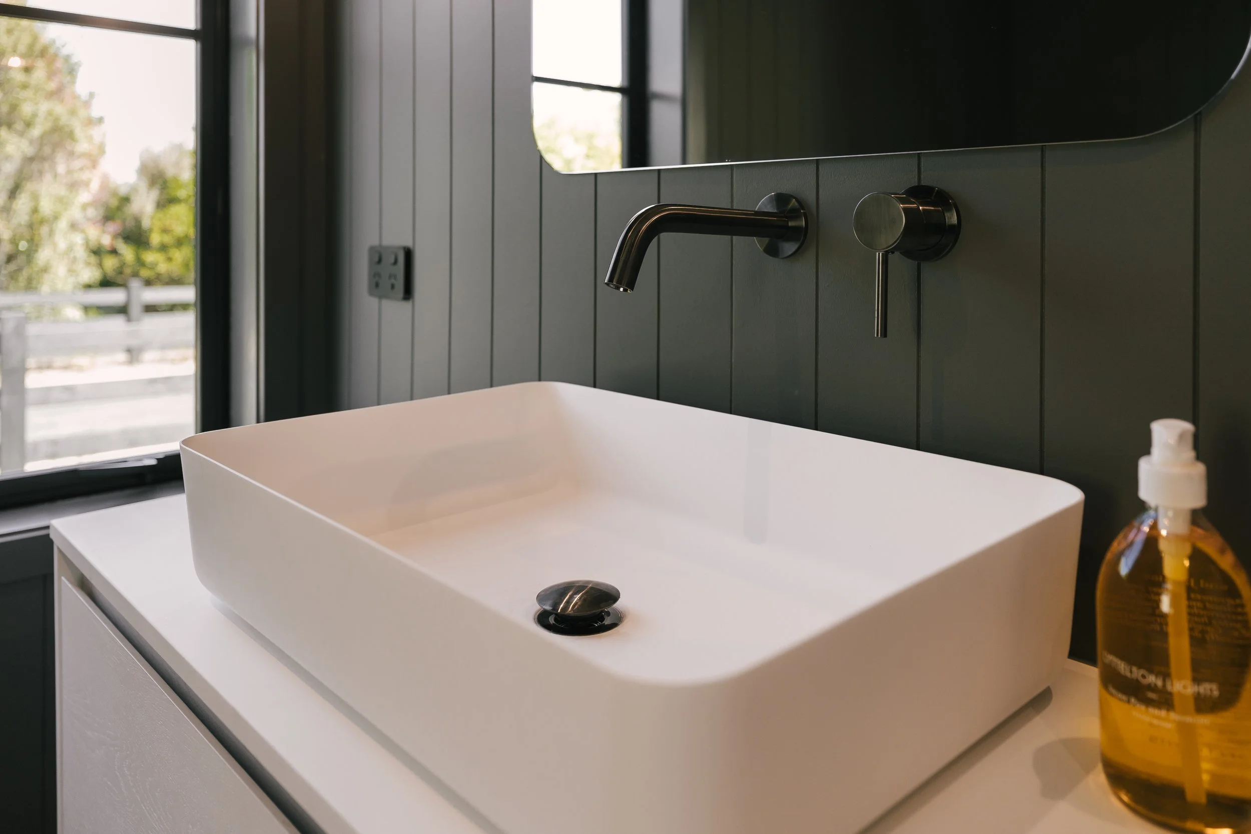 A white rectangular sink on a white vanity in front of a gray wall with a black faucet. A soap dispenser is on the right side of the sink. There is a window in the background showing trees and a cloudy sky.