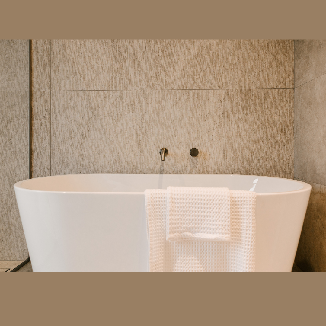 Freestanding white bathtub against beige tiled wall with a white textured towel draped over the side.